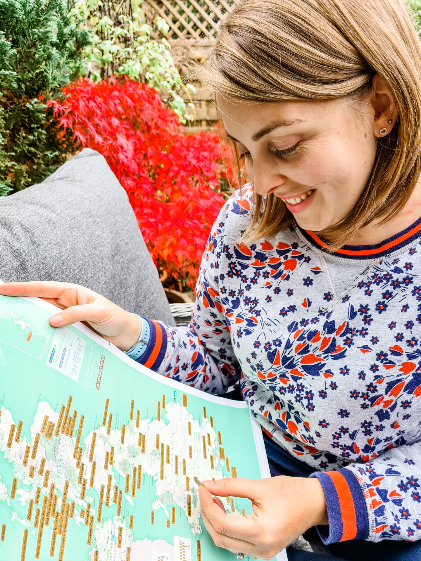 Woman smiling as she holds the Great British Outdoors scratch map on her lap with a coin in her left hand