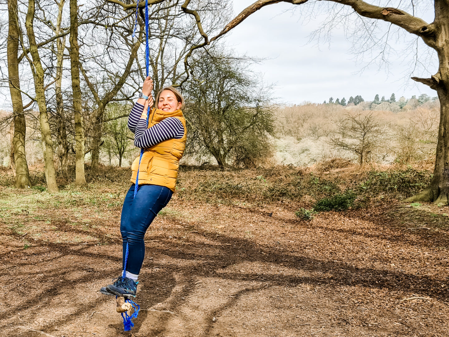 woman in yellow gilet swings on a rope swing smiling in the sunshin