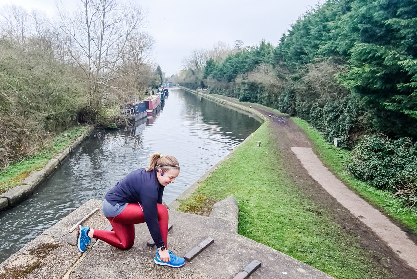 Woman in red running leggings and black top kneels to tie her shoelaces on canal towpath next to the canal (seen behind her)