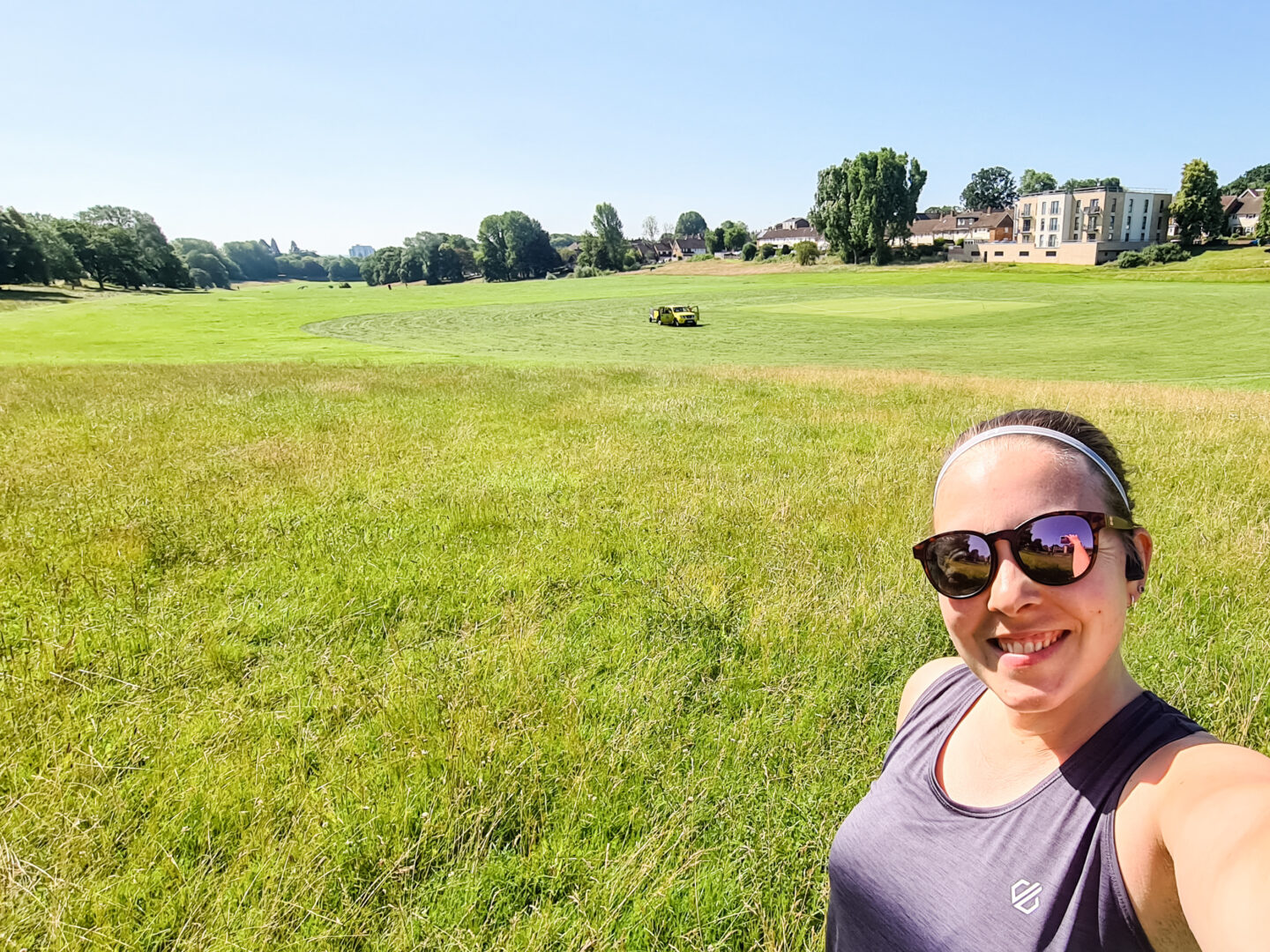 Selfie of woman in grey running vest and sunglasses, with green field behind her