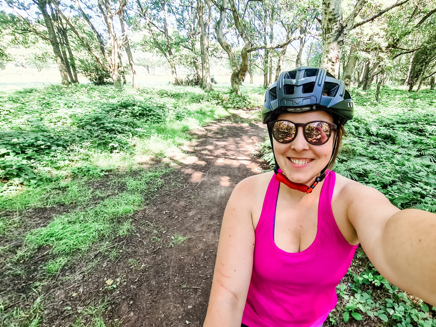 A woman in cycling kit smiles at the camera with a wood behind her, a muddy trail disappearing into the distance.