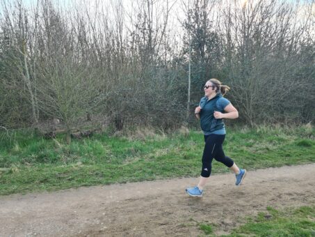 Woman in black leggings, blue running top and green gilet runs along a country trail