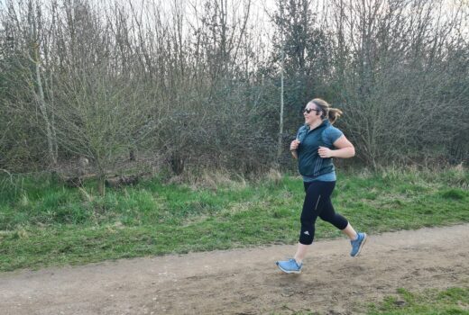 Woman in black leggings, blue running top and green gilet runs along a country trail