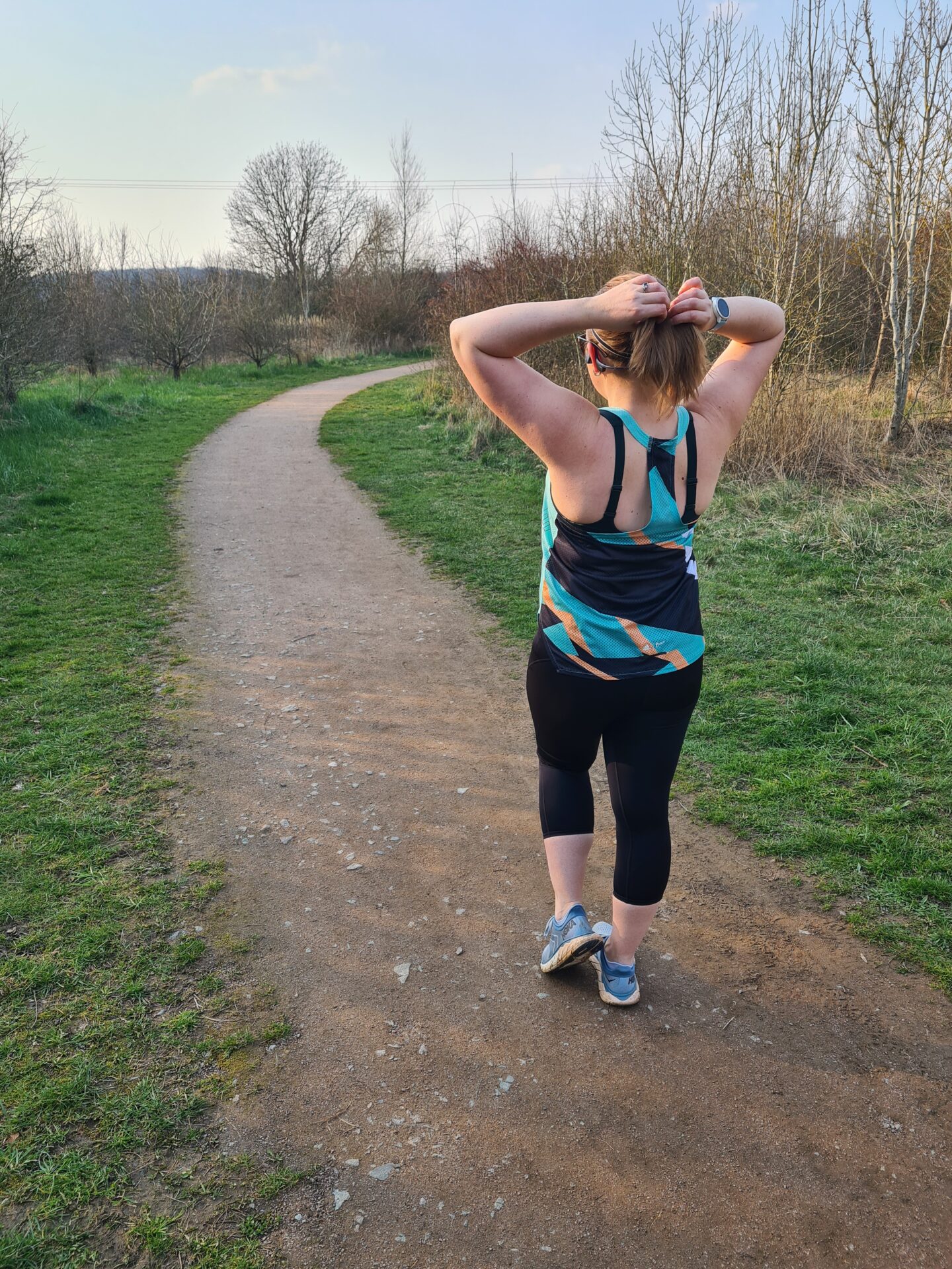 Woman stands with her back to the camera, tying up her hair. She is wearing black running leggings and a green and yellow running vest. 