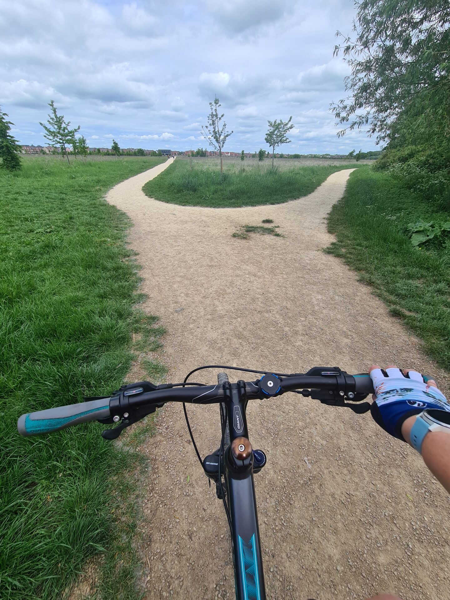 bike handlebars with one gloved hand gripping them. A dusty gravel path stretches out in front of the bike with green grass and trees around. 