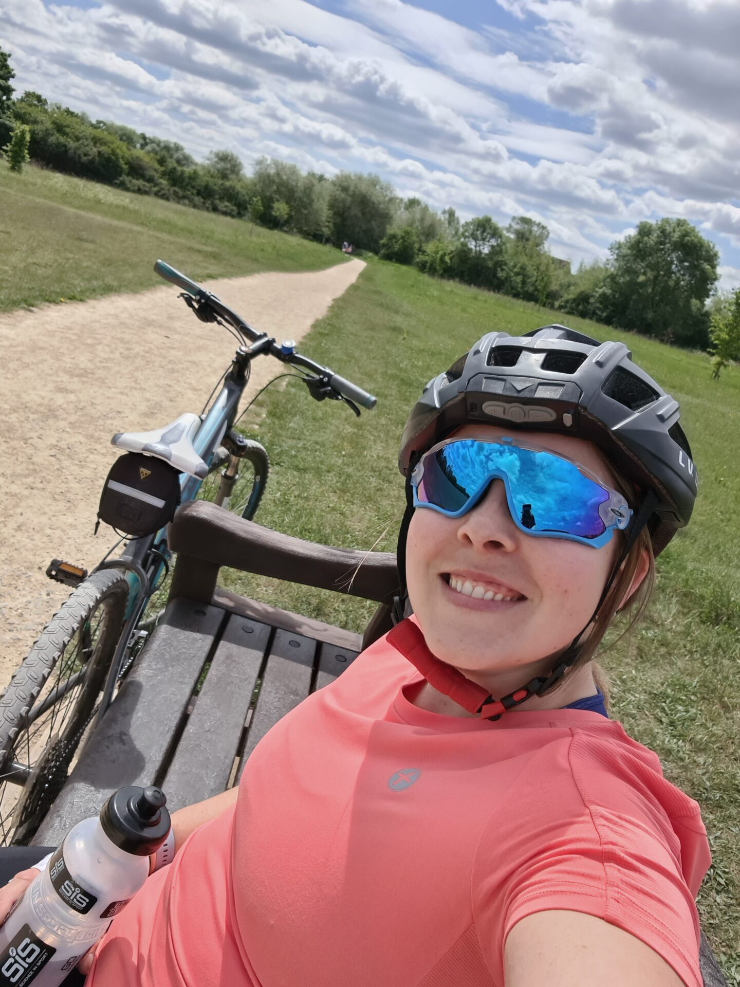 selfie of a smiling woman wearing a pink top, black cycle helmet and blue sunglasses. She is sitting on a bench with a bike next to her. 