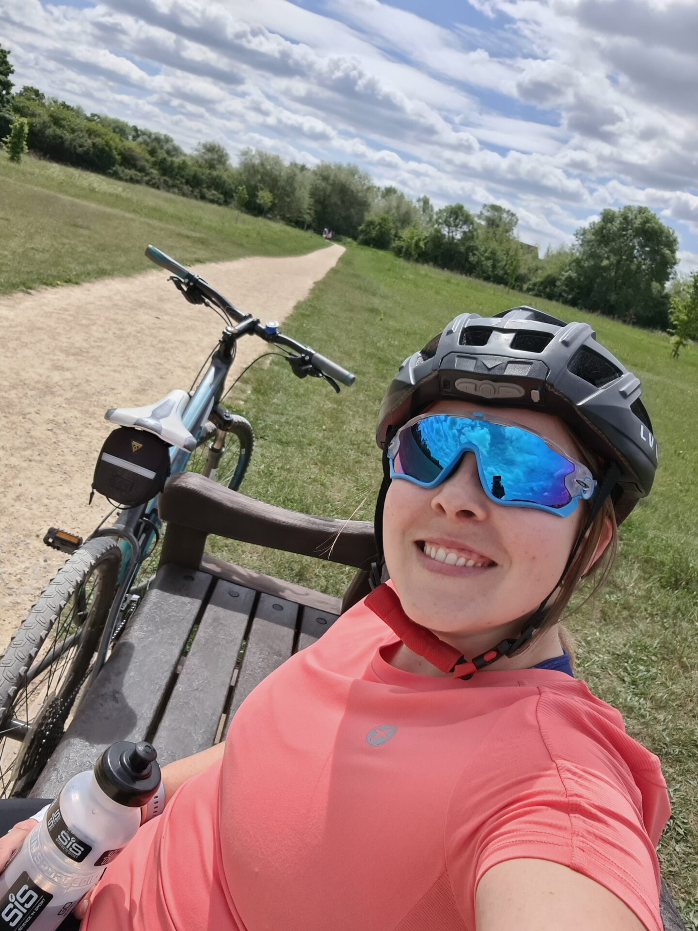 selfie of a smiling woman wearing a pink top, black cycle helmet and blue sunglasses. She is sitting on a bench with a bike next to her.