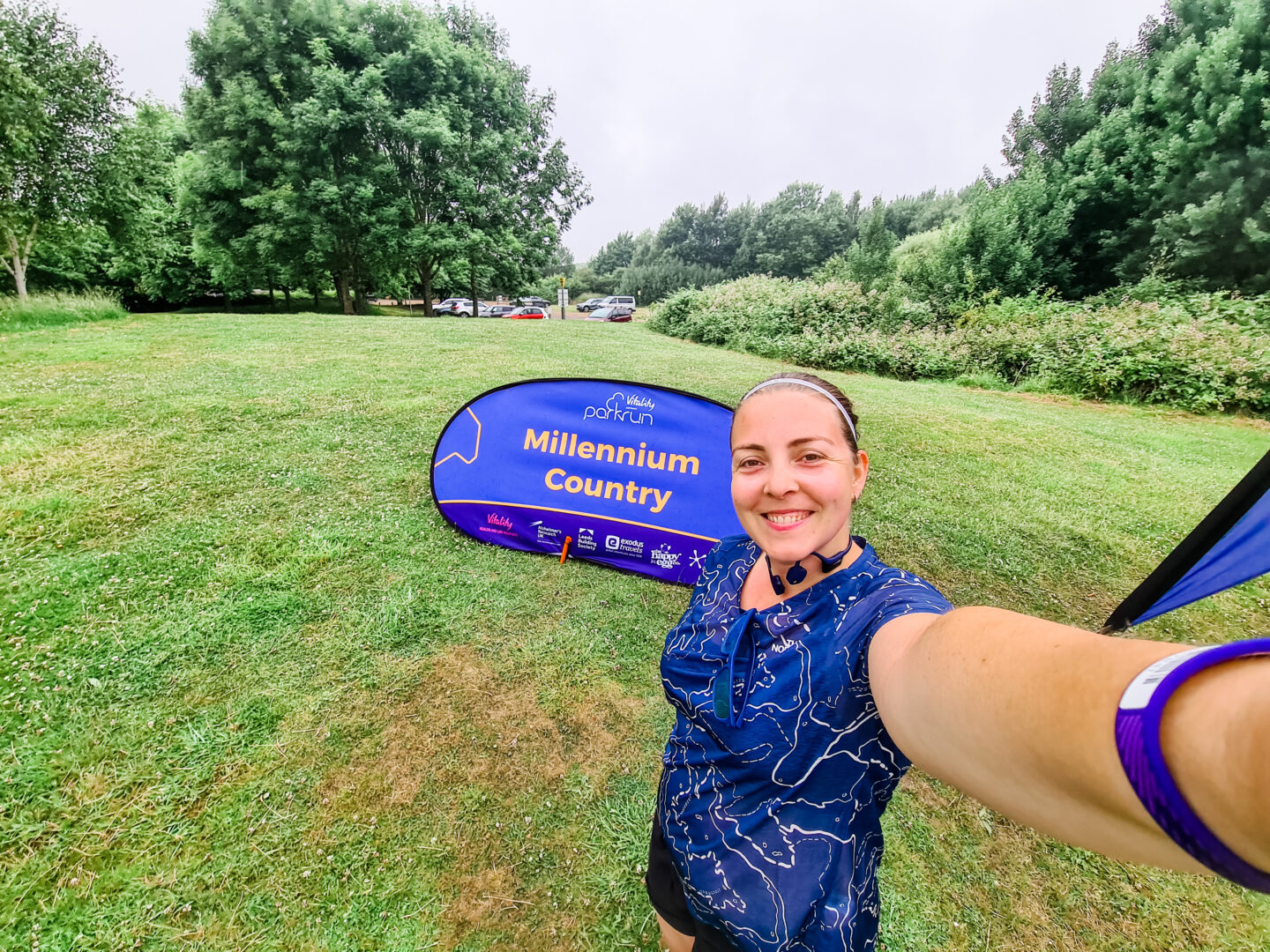 selfie of a smiling woman wearing a running top. She is standing on green grass in front of a blue sign that reads Millennium Country parkrun