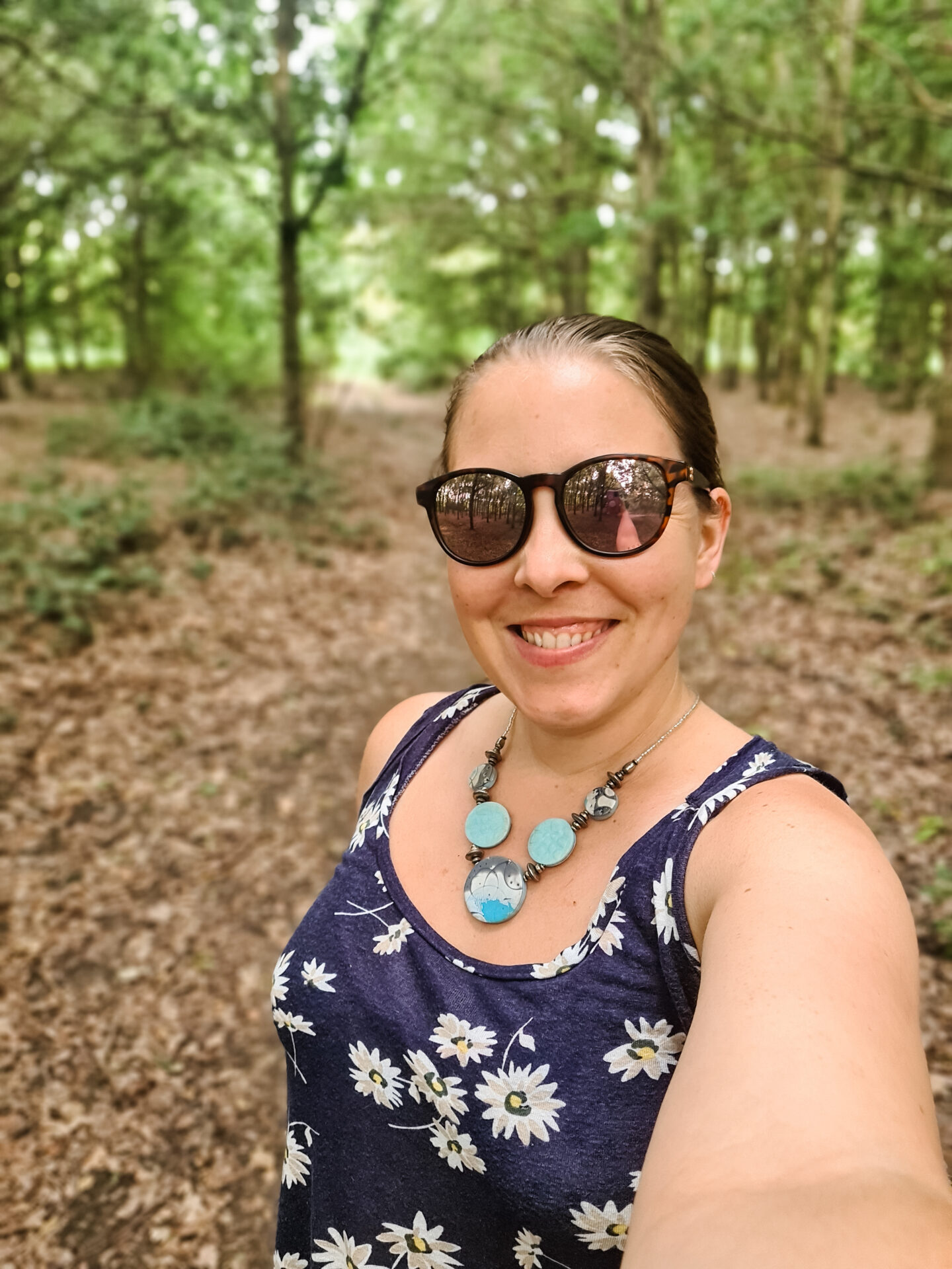 image of a woman wearing sunglasses and blue top with white daisies on it. She is smiling. Behind her is some trees in a wood