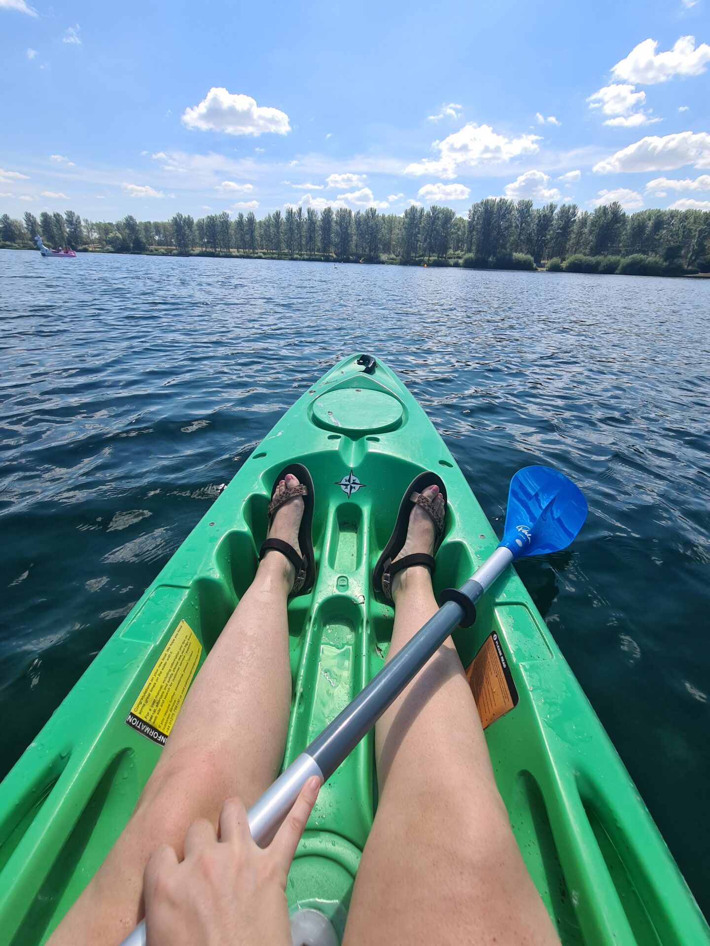 Image of legs in a kayak. A hand is holding a paddle. The water is blue with a row of trees in the background
