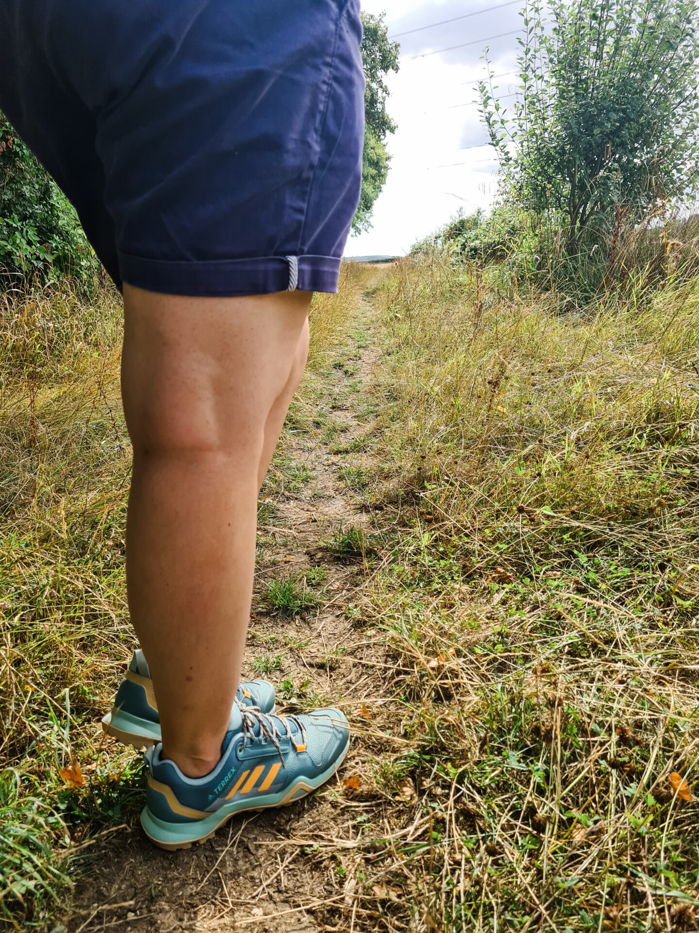 Image of legs, wearing blue shorts and green and orange adidas hiking shoes. Standing on a country path with grass around. 