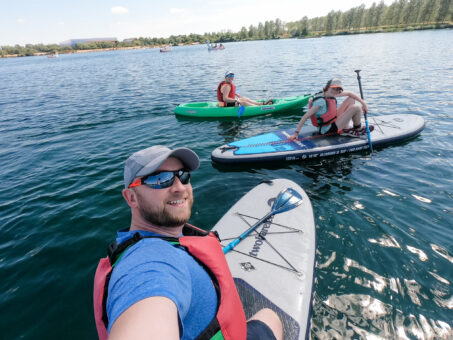 family photo with a man on a paddleboard, girl on a paddleboard and woman sitting in a green kayak. the water is blue with a row of trees in the background