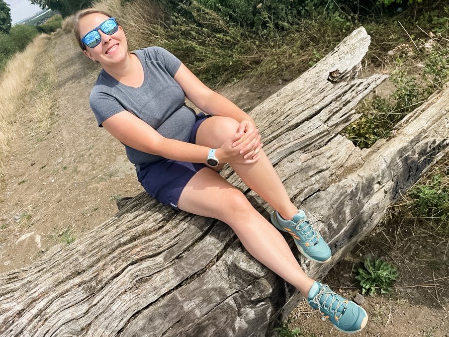 image of a woman sitting on a log. She is wearing a grey tshirt, blue shorts and green and orange adidas hiking shoes. She is wearing blue sunglasses and smiling. 
