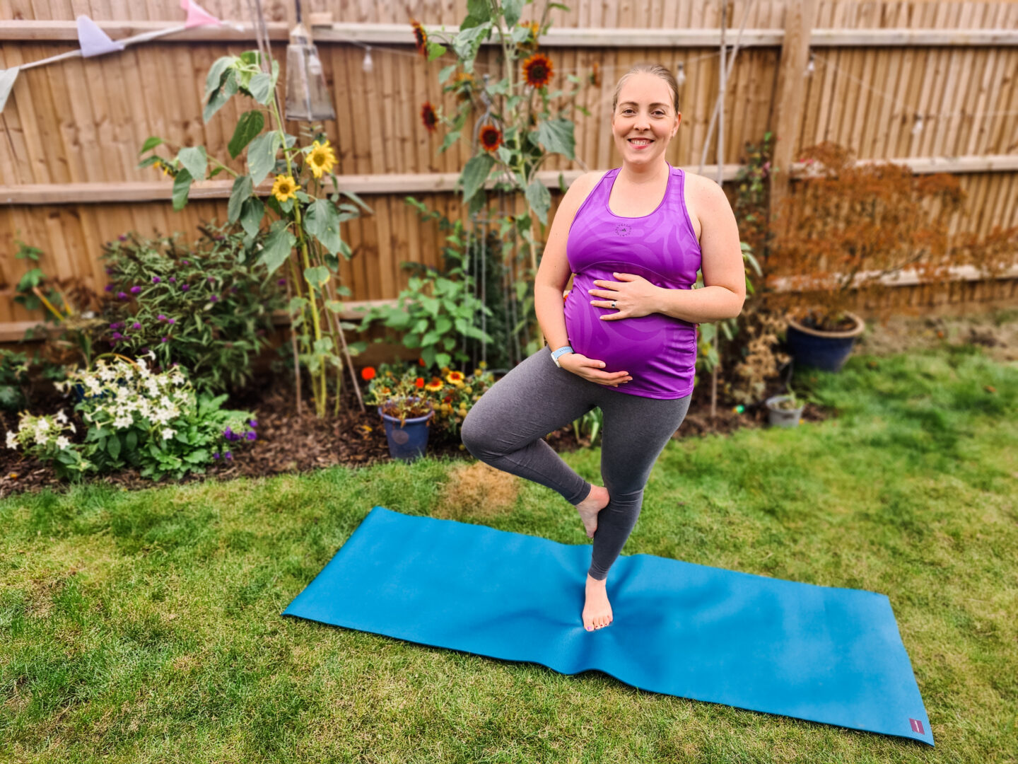Pregnant woman standing on a yoga mat doing tree pose. She has her hands on her belly, and is wearing grey leggings and purple tank top. Behind her is a blurred background of grass and plants.