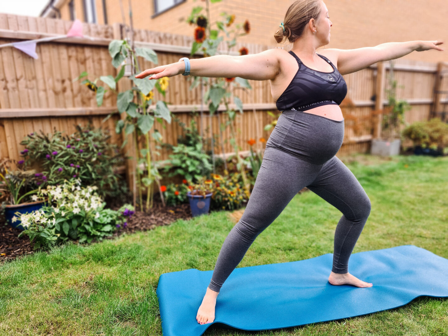 Pregnant woman wearing grey leggings and black sports bra is standing on a yoga mat doing Warrior pose. Behind her is blurred background of grass and plants