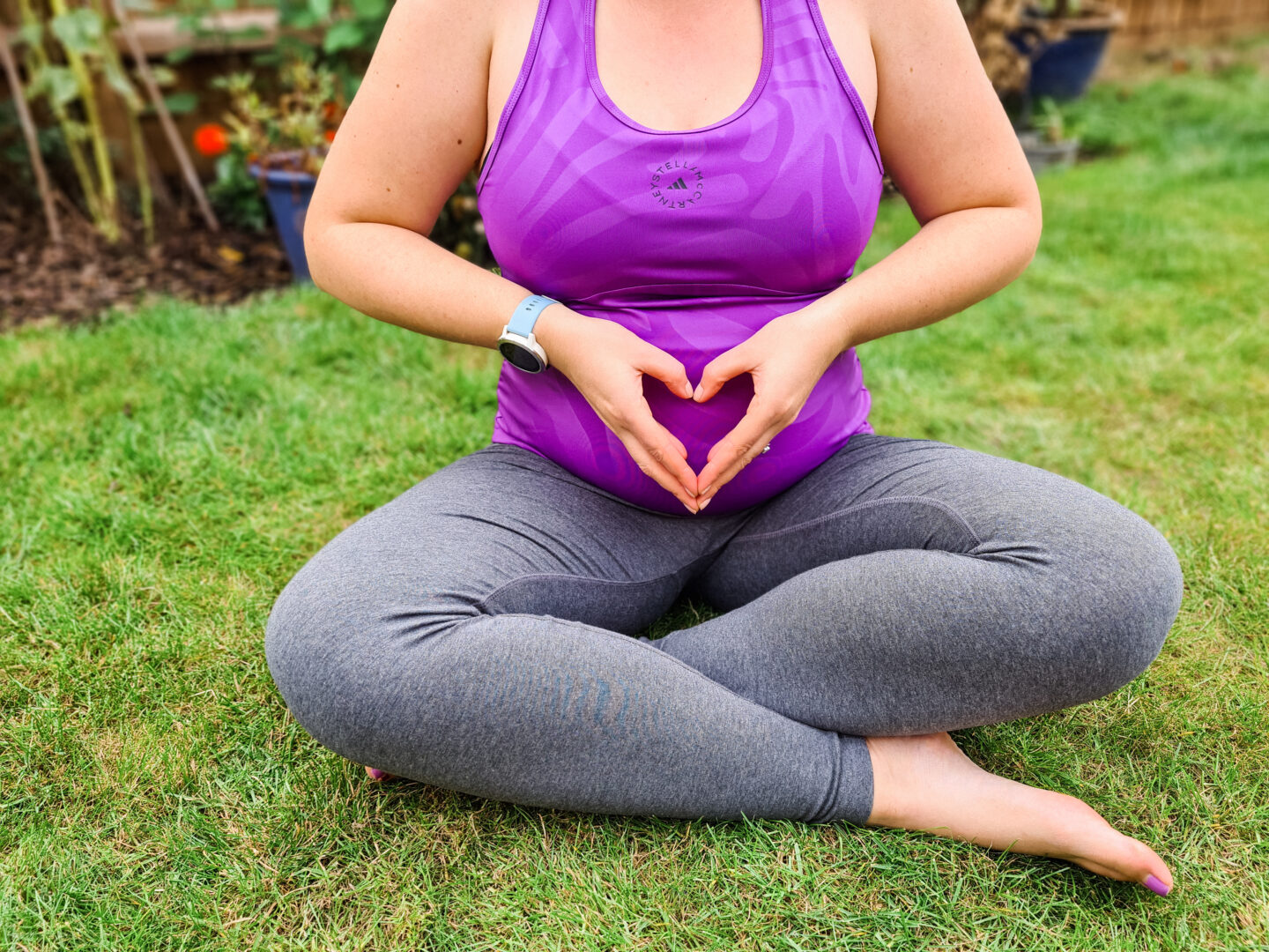 Pregnant women sits cross-legged on the grass, with her hands making a heart on her belly. She wears grey leggings and a purple top. 