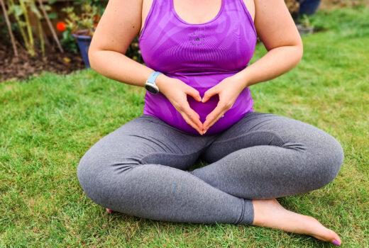 Pregnant women sits cross-legged on the grass, with her hands making a heart on her belly. She wears grey leggings and a purple top.