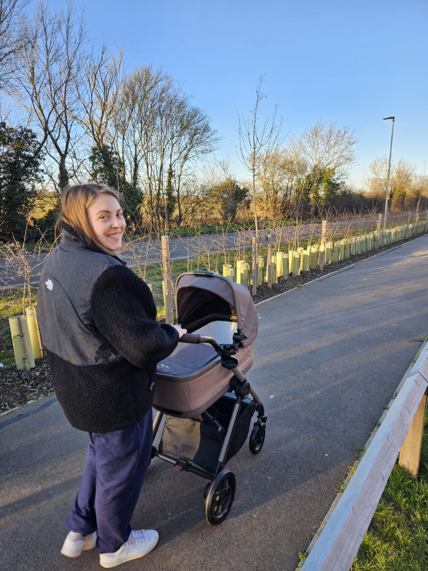 Woman wearing a black fleece and blue jogging bottoms is pushing a pram along a pavement lined with trees. Her head is turned back to look at the camera and she is smiling. 