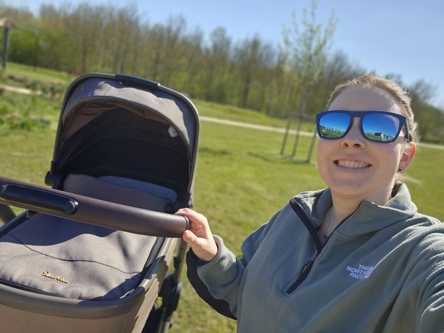 Woman wearing sunglasses is smiling at the camera with her hand resting on the handle of a pram. The background is blurred and shows green grass and trees.