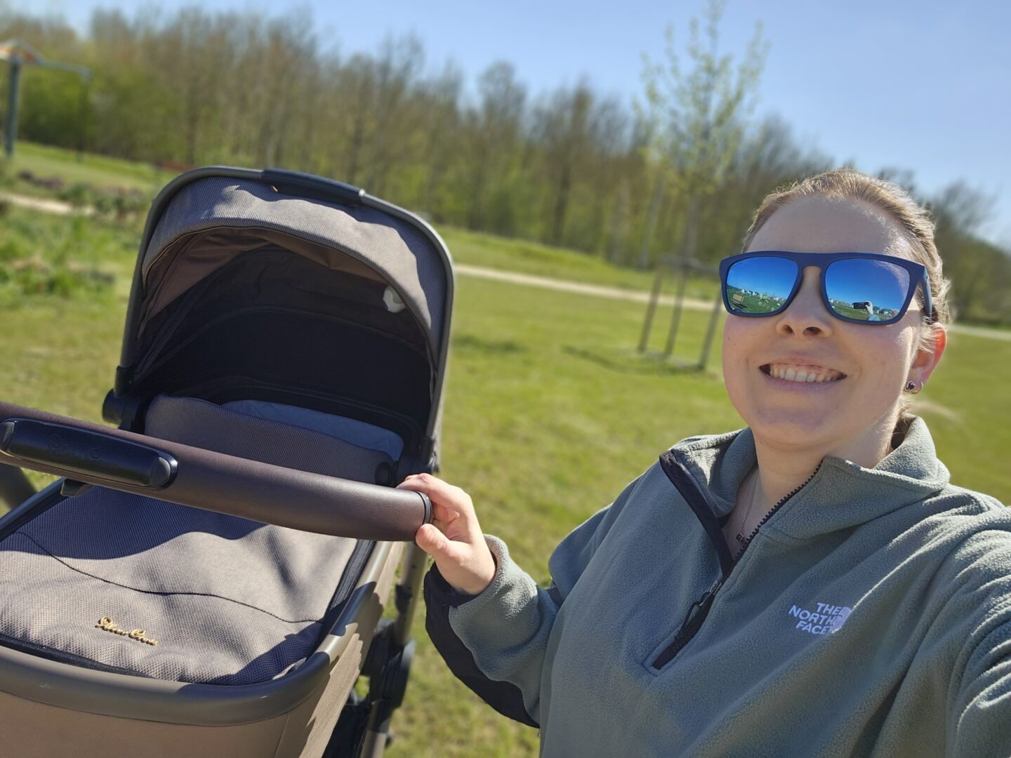 Woman wearing sunglasses is smiling at the camera with her hand resting on the handle of a pram. The background is blurred and shows green grass and trees. 