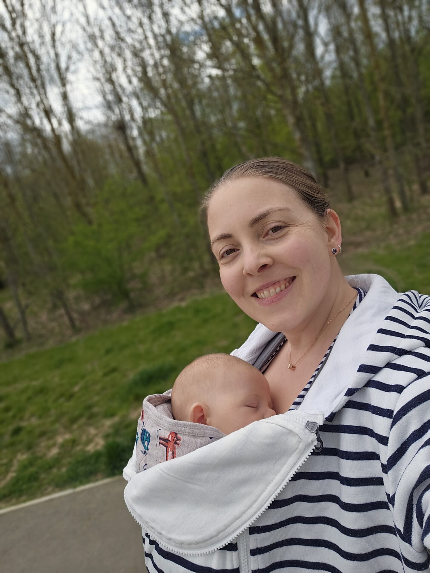 Woman wearing a striped blue and white hoody is carrying a baby on her chest. The baby is sleeping and the woman is smiling. The background is blurred and shows green grass and trees.