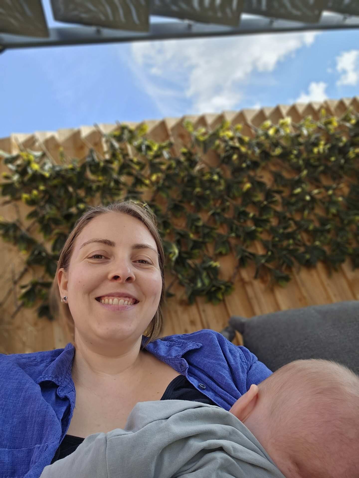 woman sitting outside with blue skies behind her. she smiles at the camera and is holding a baby who is breastfeeding 
