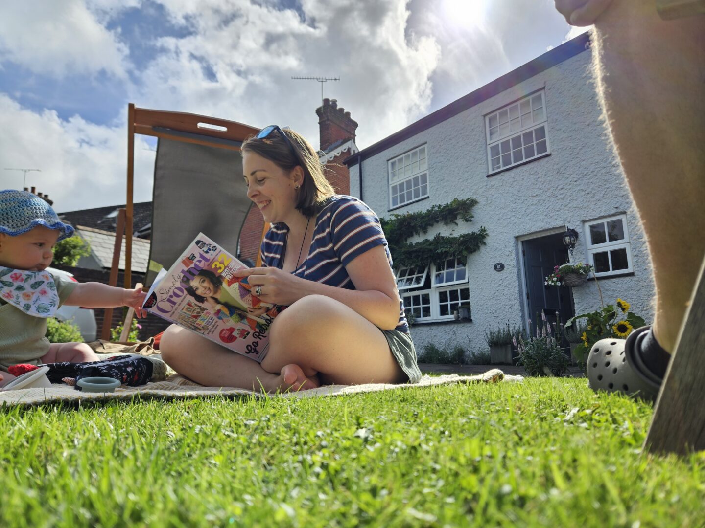 woman in striped top sitting on grass reading crochet magazine, with white cottage behind her