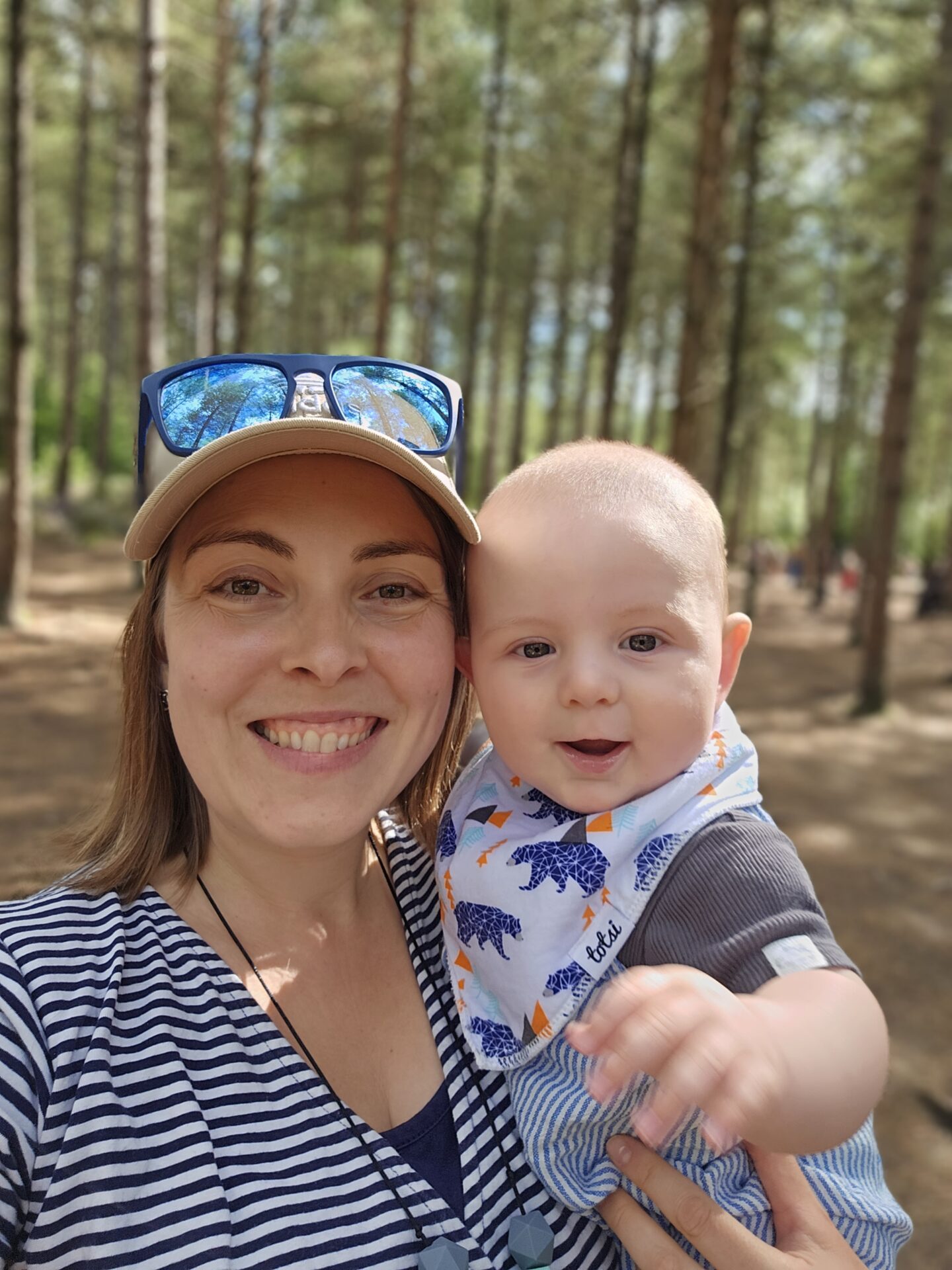 woman wearing a striped top and baseball cap is smiling at the camera. she is holding a baby in her arms who is  also smiling. the background is blurred but shows trees.