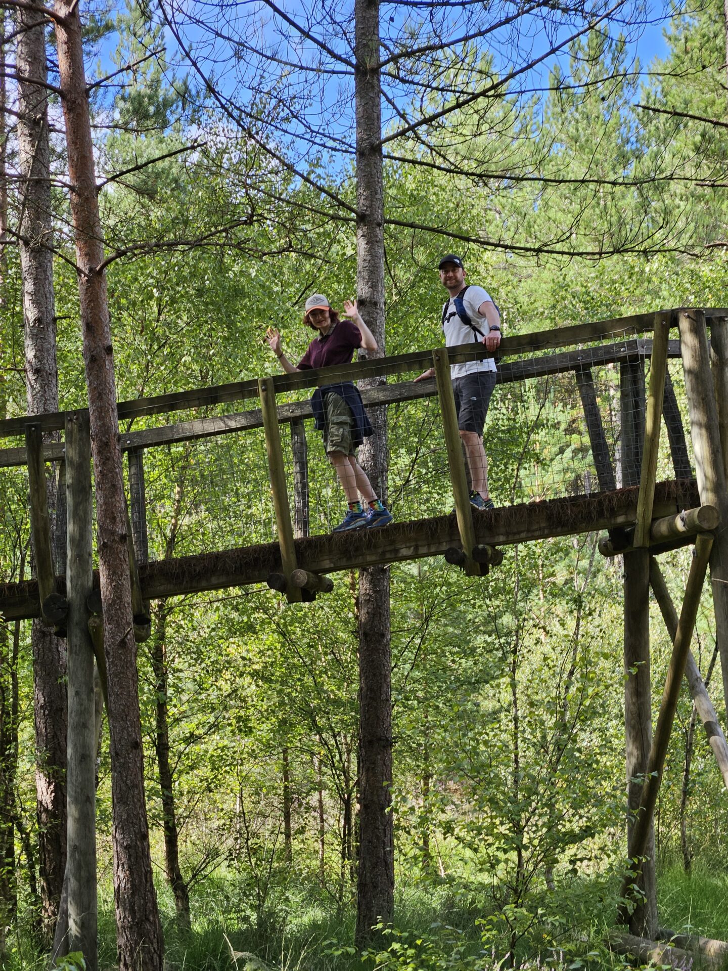 man and young woman standing on an aerial wooden bridge in a forest, both waving at the camera. 