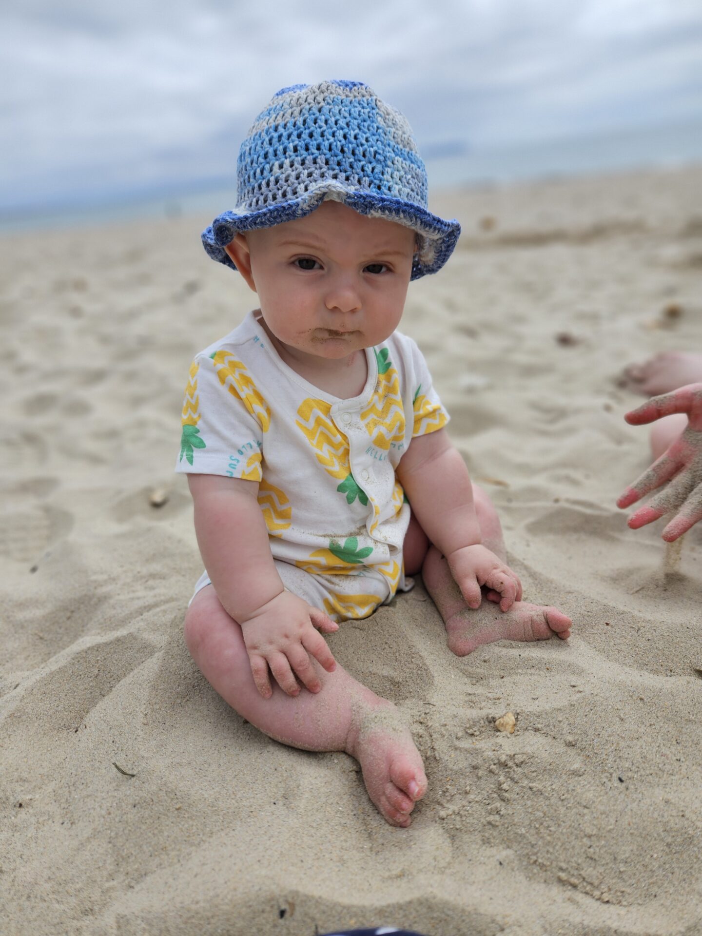 A small baby in white romper and blue crochet hat sits on a sandy beach