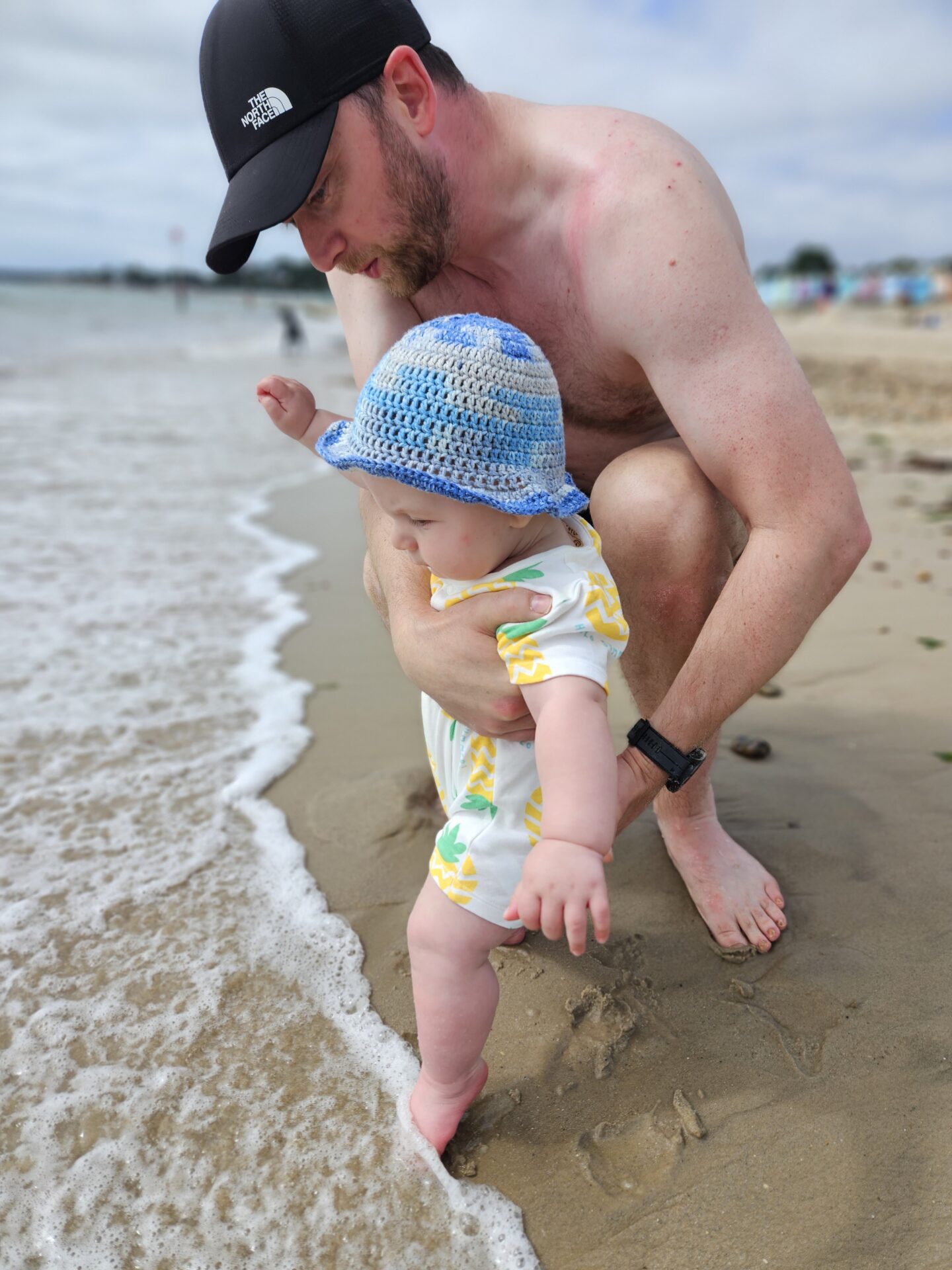 man in swim shorts and baseball cap crouches on a sandy beach holding a small baby as he dips his toes into the sea