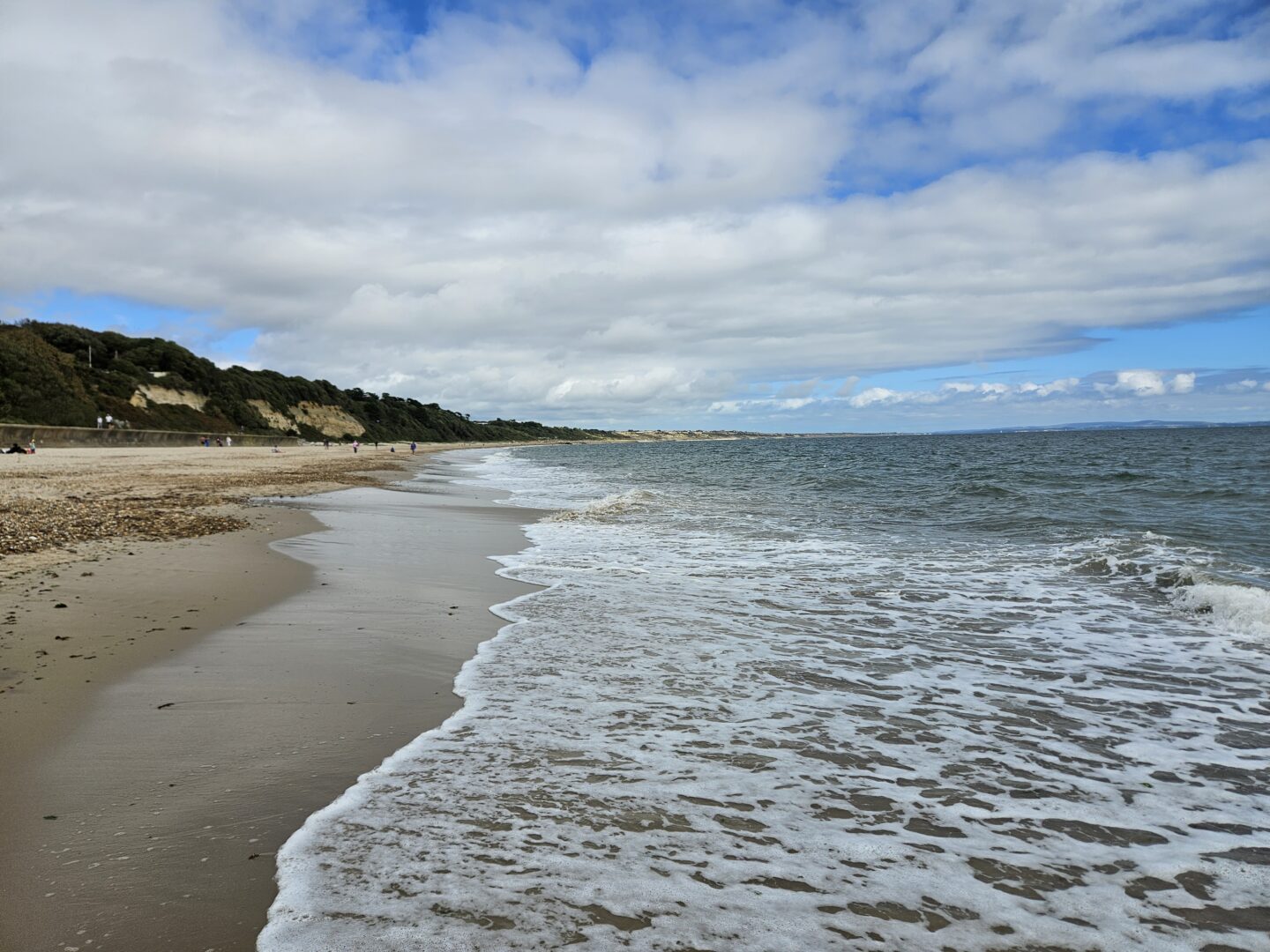 landscape photo of a UK beach with blue grey sea with white foam, sand, and blue skies with white clouds. 