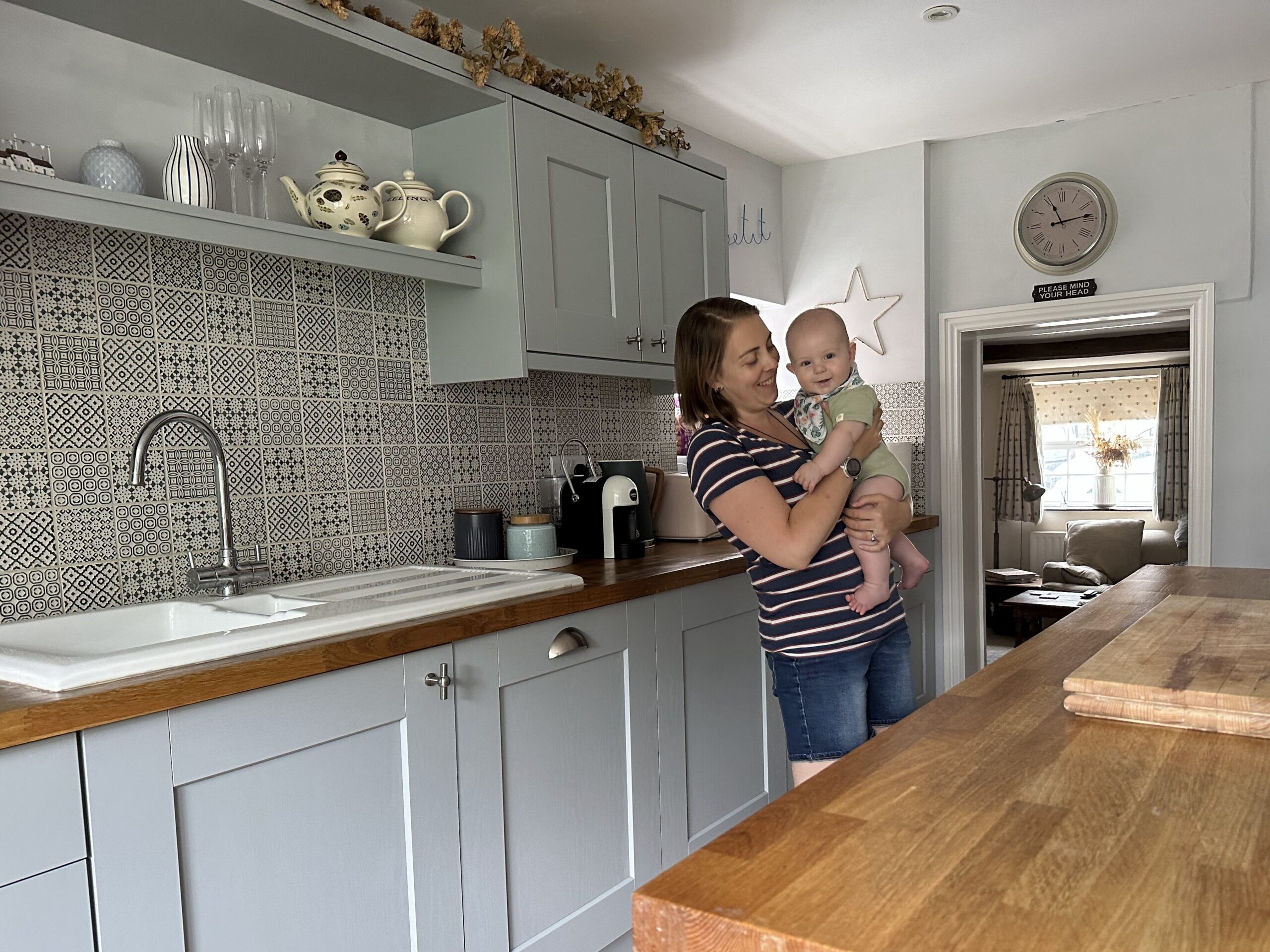 woman in striped top holding a baby in a farmhouse kitchen with grey cupboards and wooden worktops