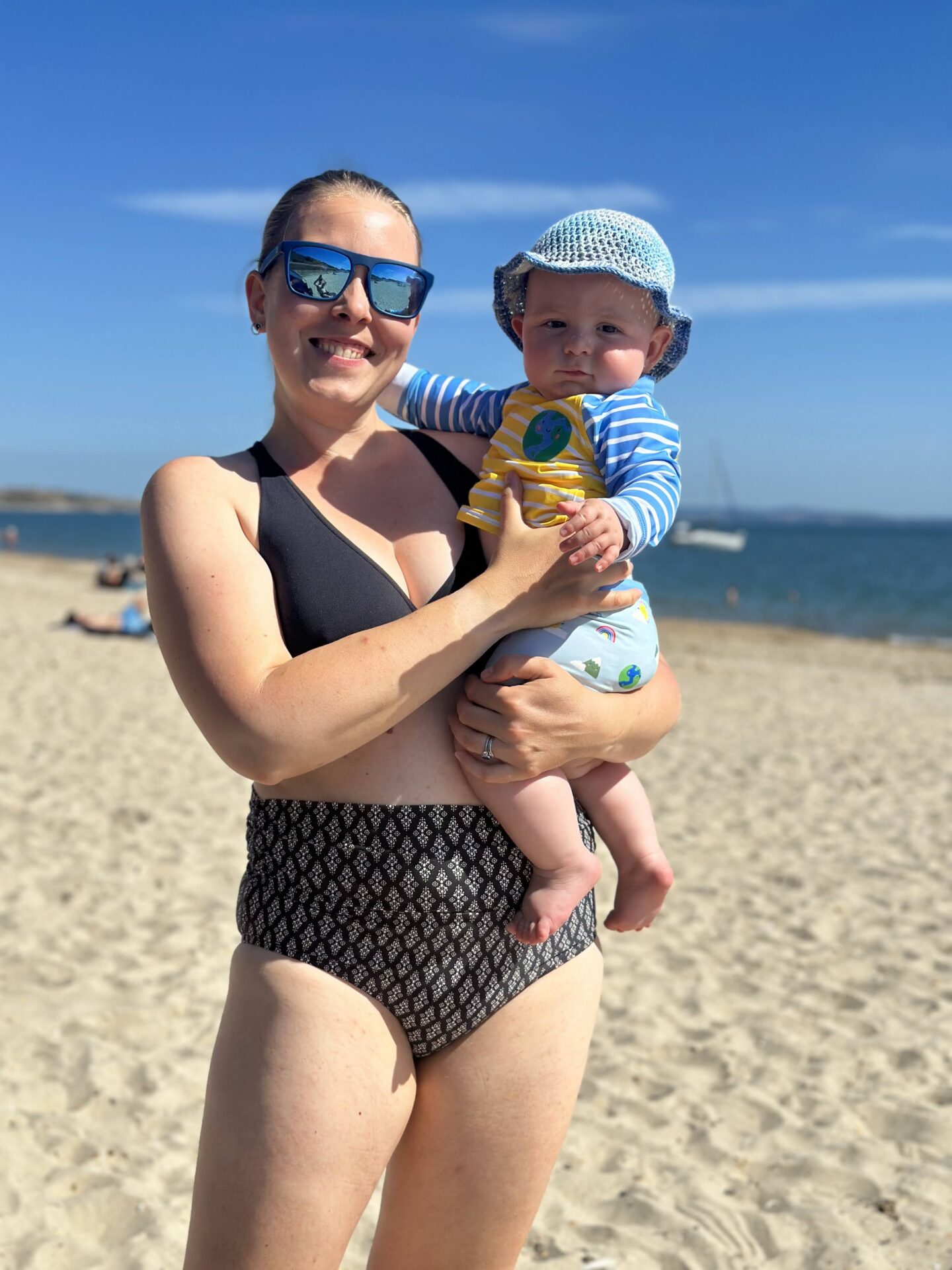 woman in a black bikini is smiling at the camera holding a baby in a striped swim outfit and crocheted hat. A sandy beach is blurred behind them. 