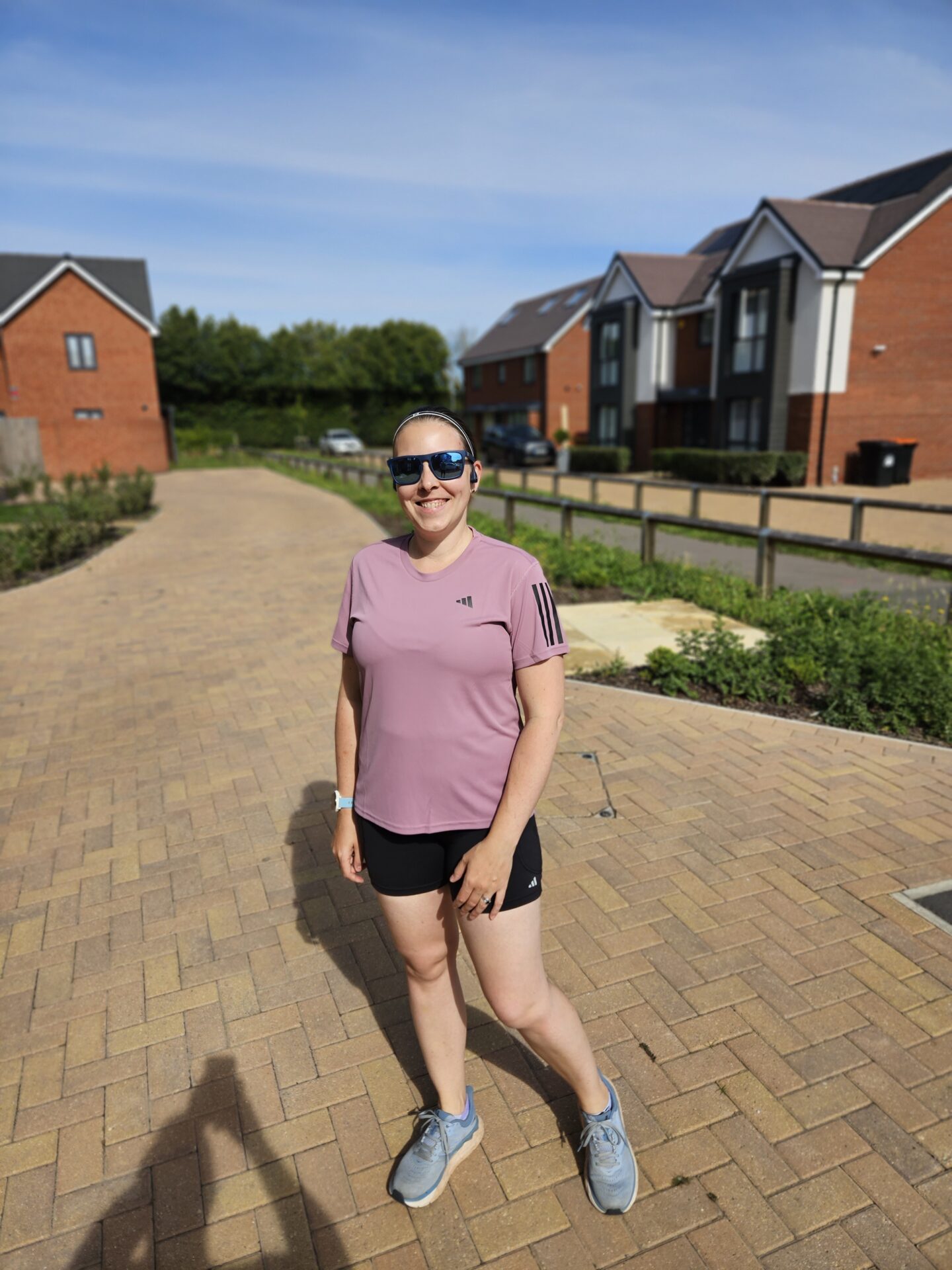 Woman in purple running top and black shorts stands on a block paved driveway in front of houses