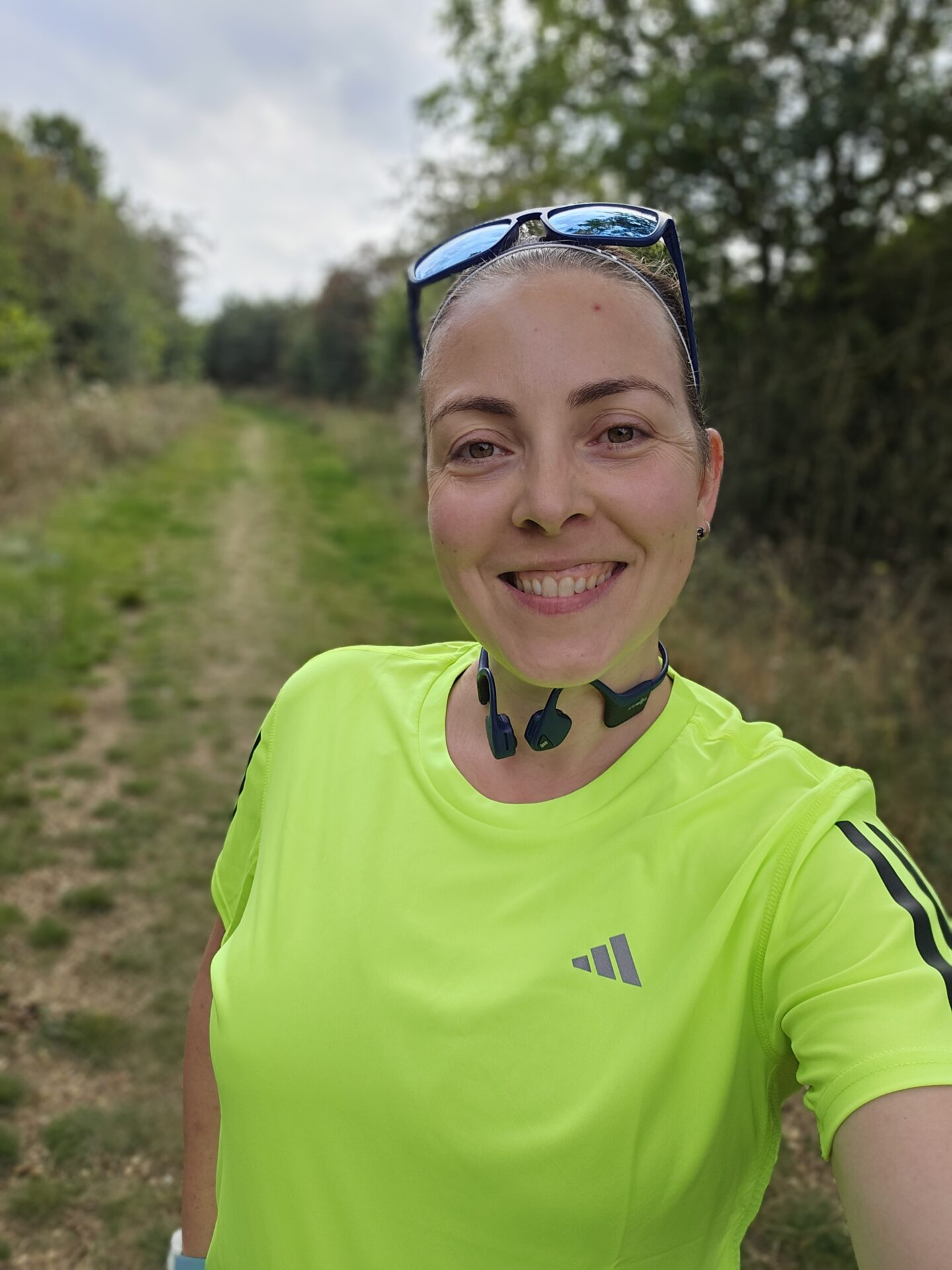 woman in fluorescent yellow running top smiles at the camera. the background behind her shows a blurred green running trail. 