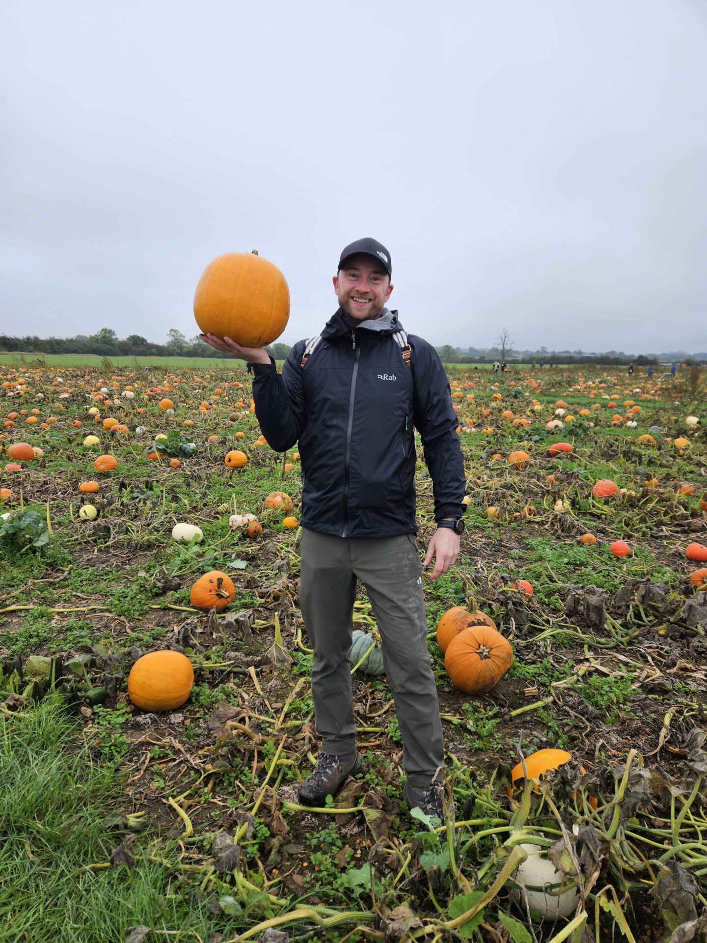 man in black coat stands smiling at the camera with a orange pumpkin in his hand
