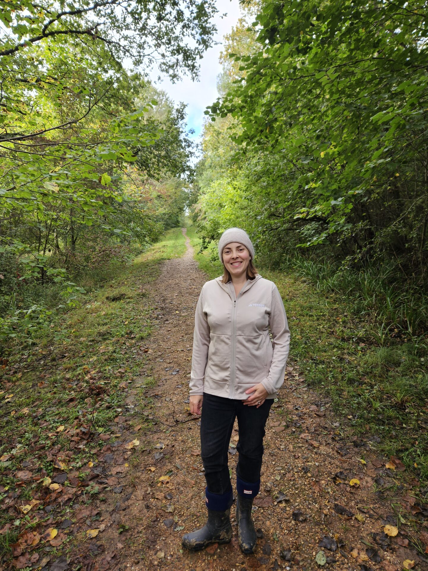 Woman wearing black jeans, wellies and a beige zip up jumper stands on a muddy path in the woods smiling at the camera. 