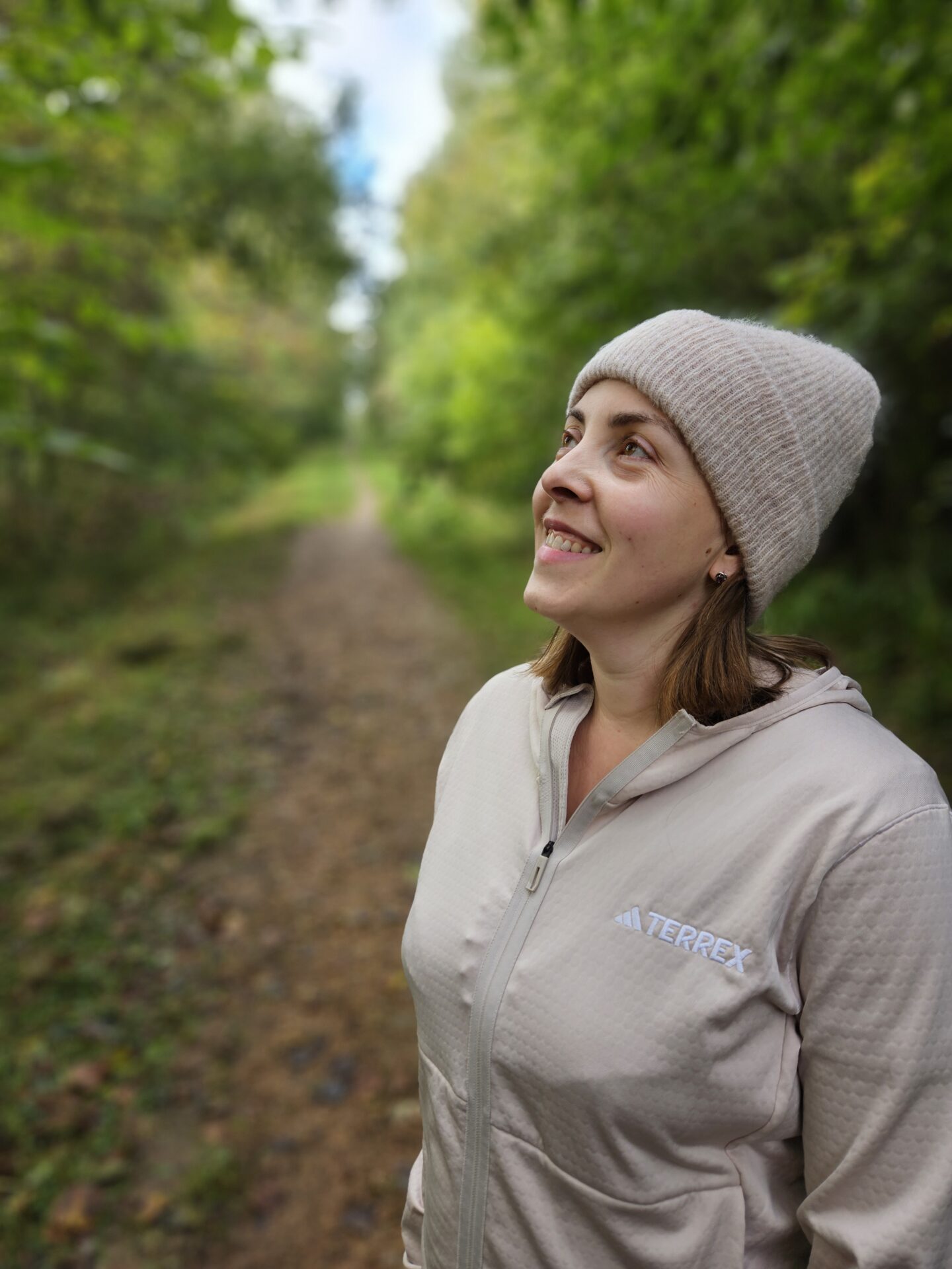Woman in beige zip up jumper and woolly hat stands smiling up to the left