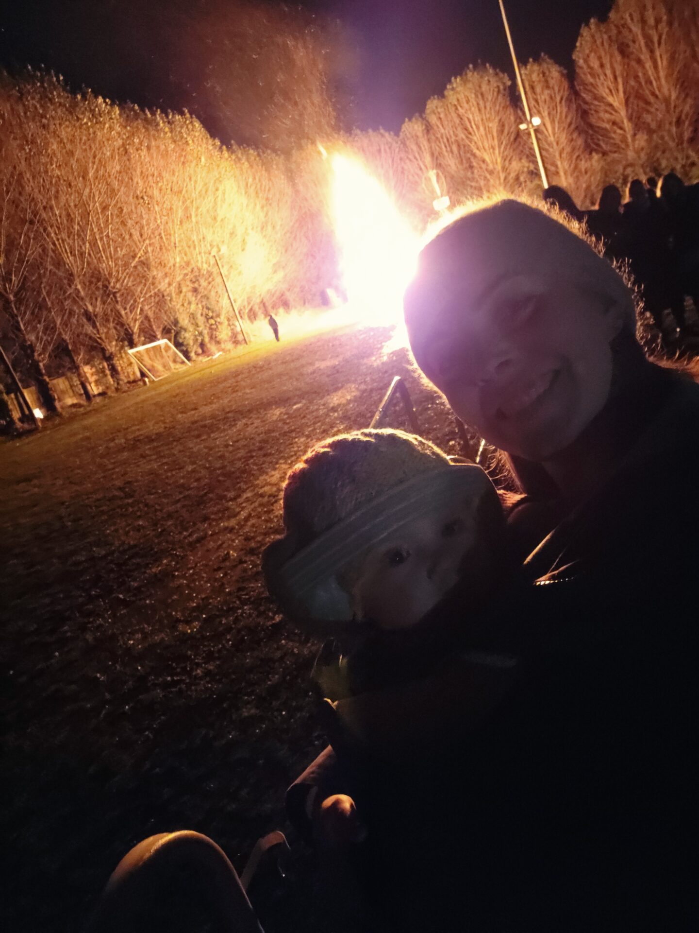Night time image showing a woman carrying a baby in a sling facing the camera. Their faces are dark and behind them is a large glowing bonfire. 
