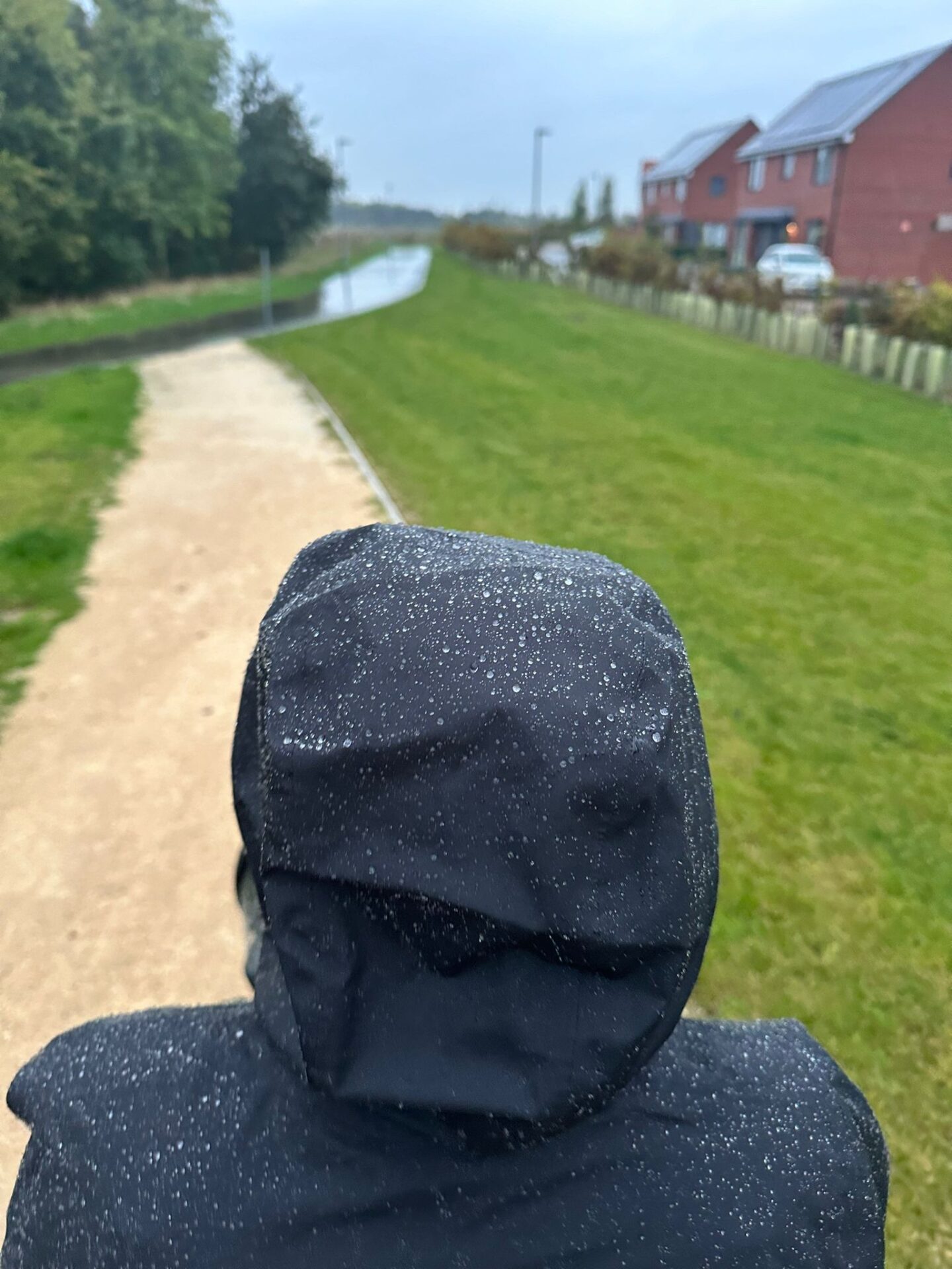Back of a person's head in a black waterproof coat. Their hood is up and you can see the rain beading on the jacket. 