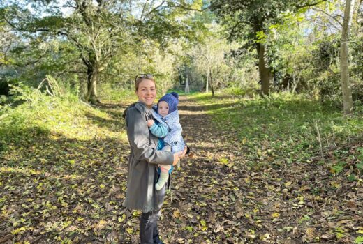 woman in green coat is carrying a baby in a sling. she is smiling at the camera and the baby is wearing a blue woolly hat. They are on a muddy path through the woods with trees behind them.