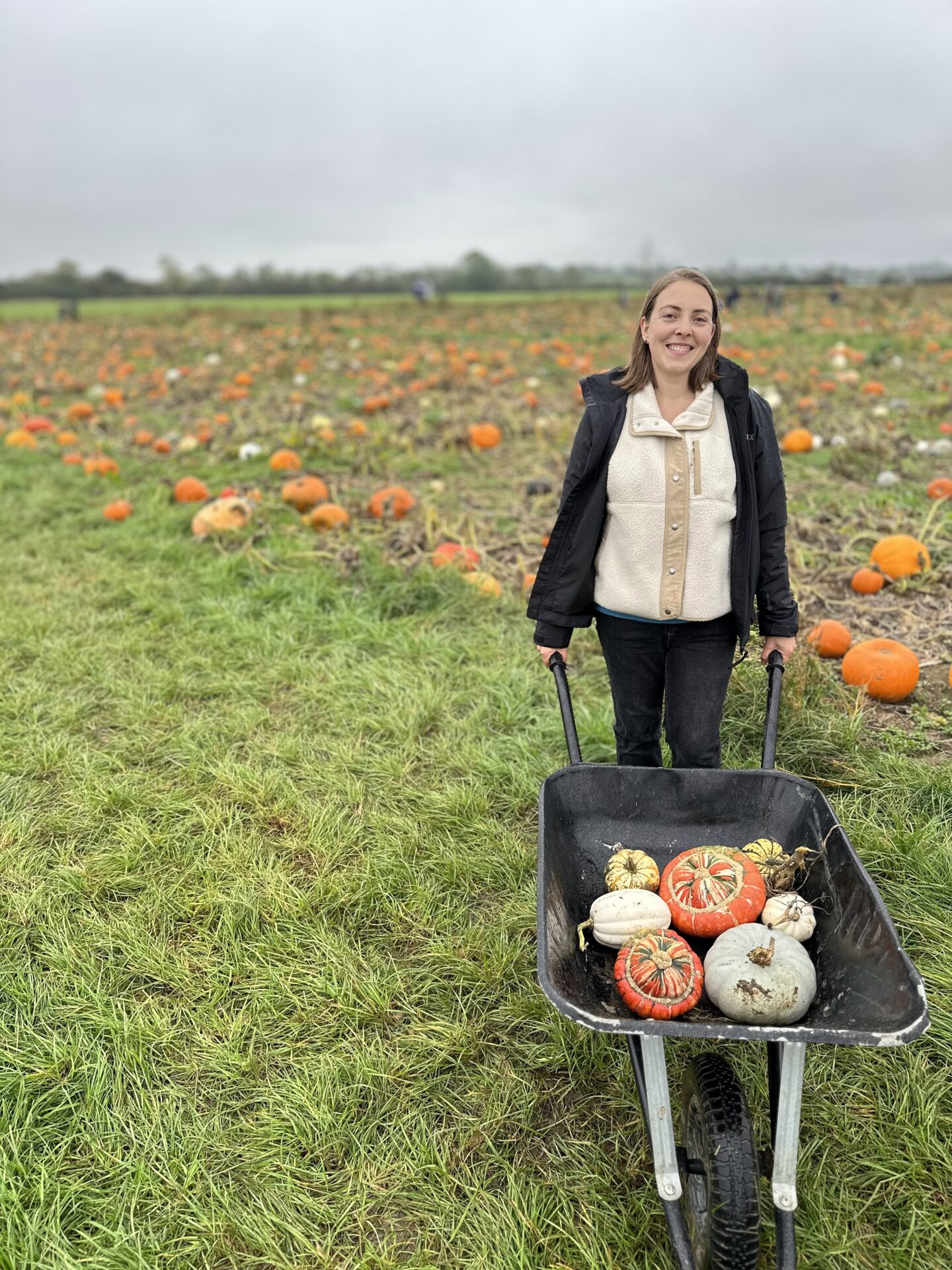 woman in a white fleece and black coat stands smiling at the camera pushing a wheelbarrow full of pumpkins 