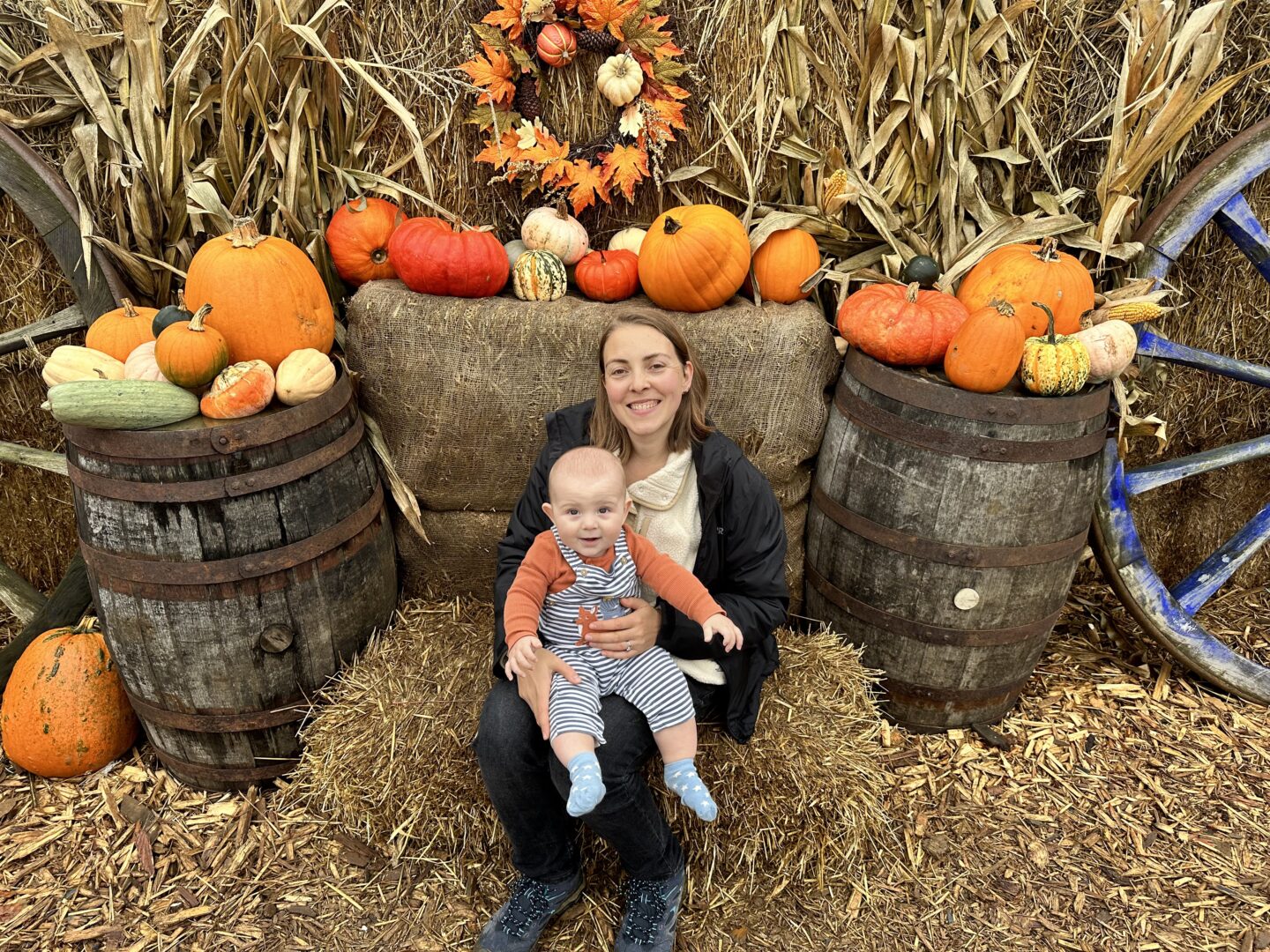 woman in a white fleece and black coat sits on a hay bale with a baby in stripy dungarees on her lap. Behind them is a display of lots of orange pumpkins