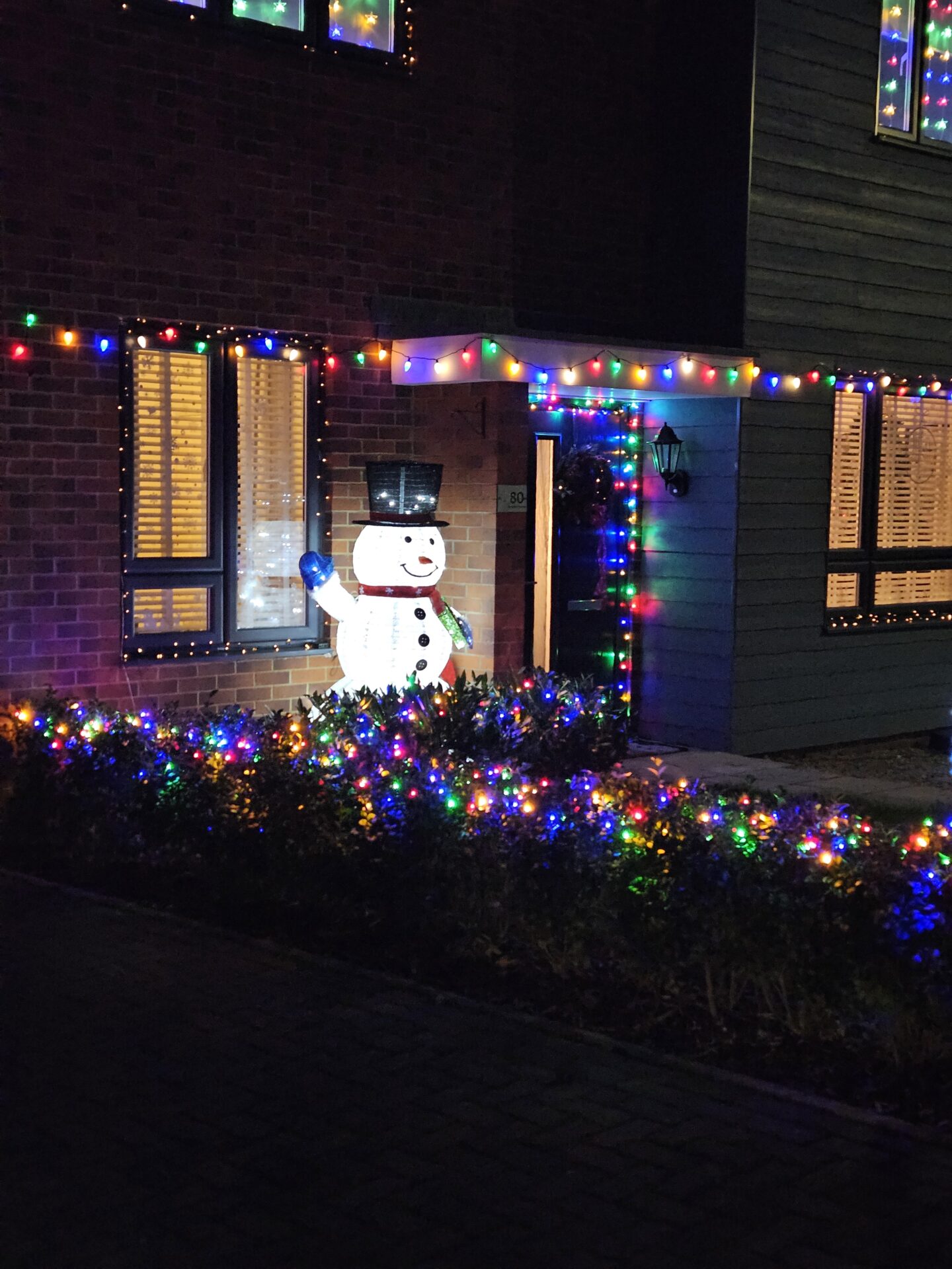 Inflatable snowman stands in front of a house which is decorated with multi-coloured Christmas lights. 