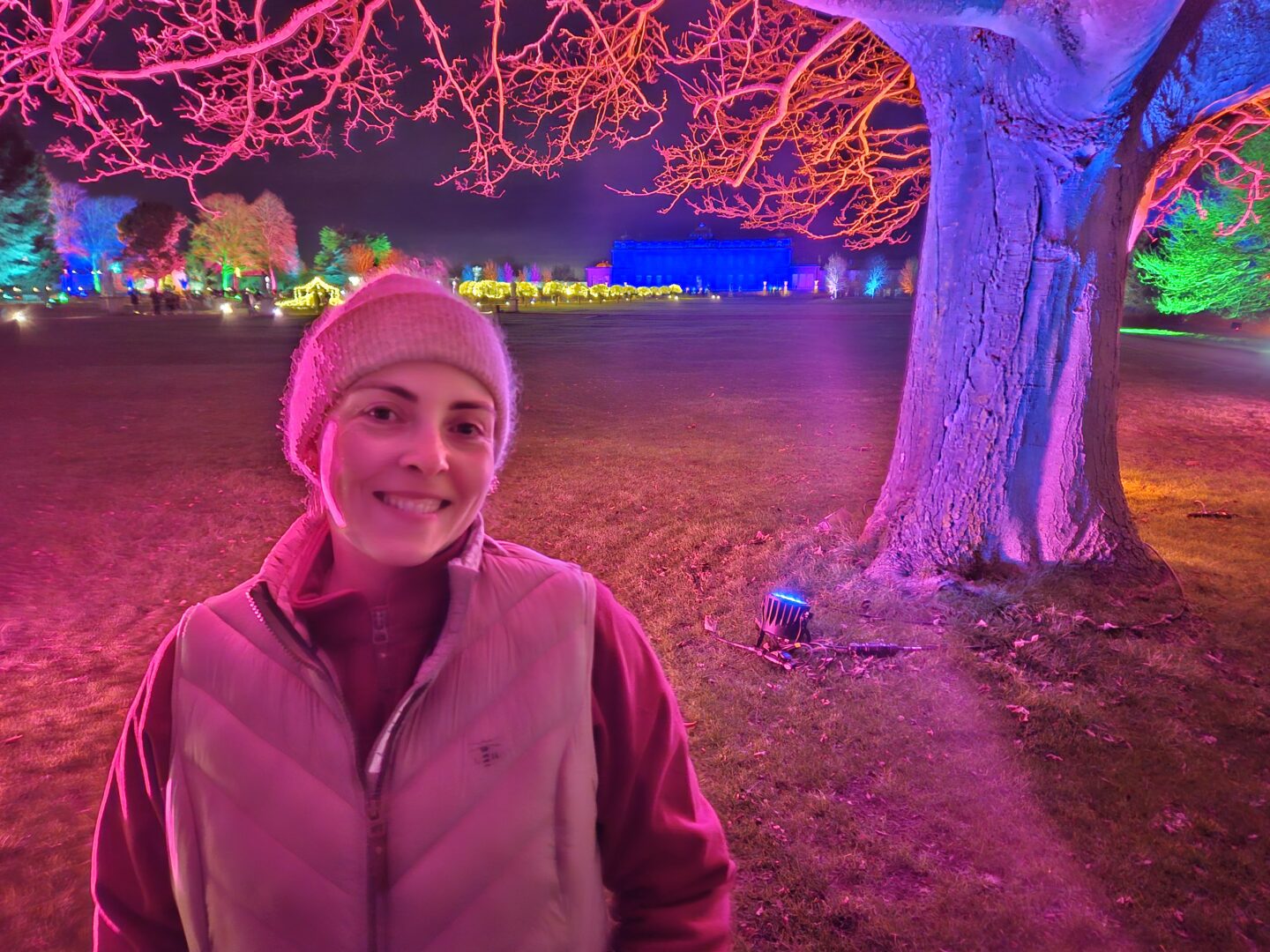 Woman in a fleece, gilet and beanie standing by a tree with lots of colourful Christmas lights behind her