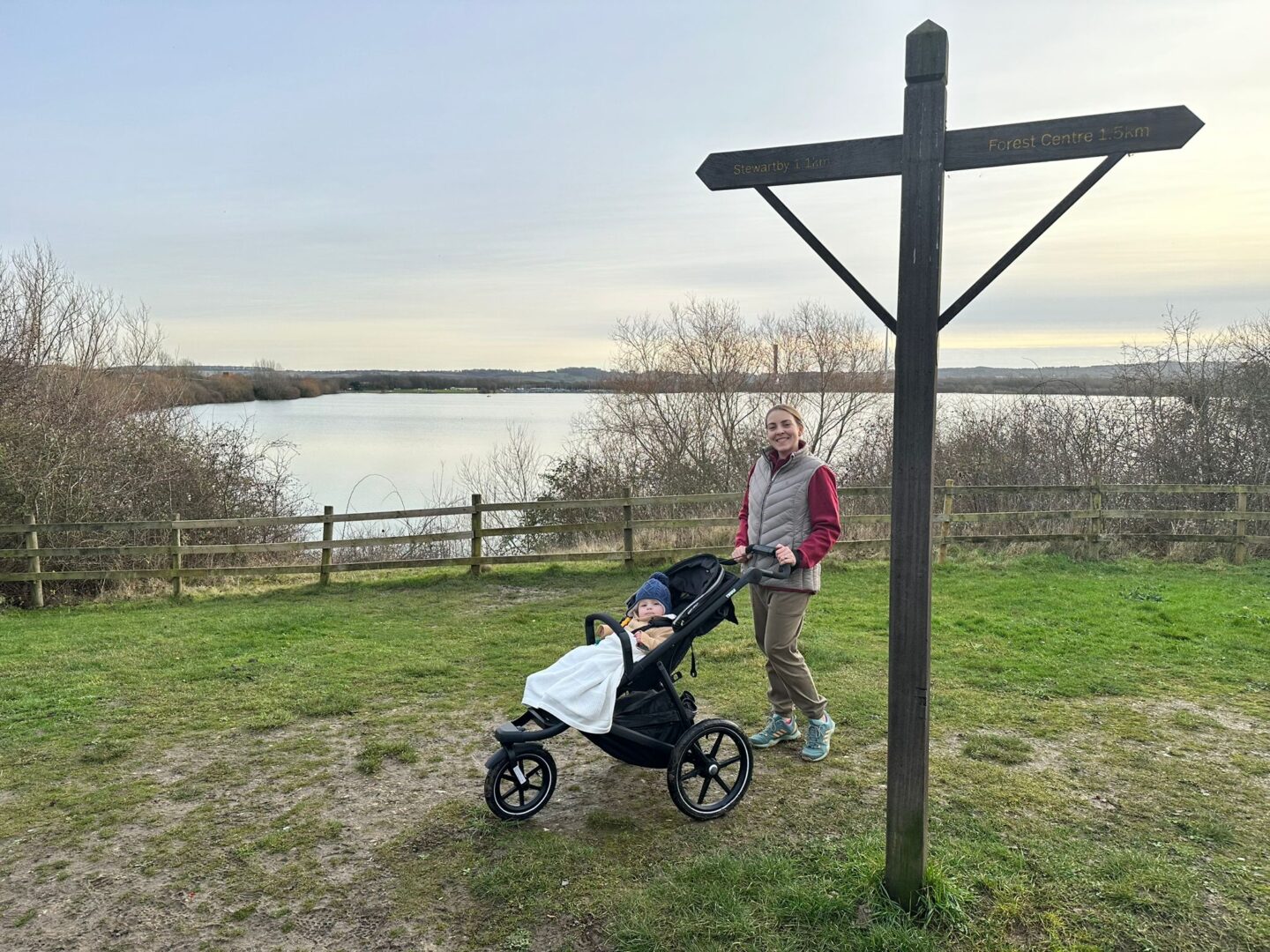 Woman pushing an off-road buggy with a baby in it stands on the grass by a wooden sign. Behind them is a lake. 