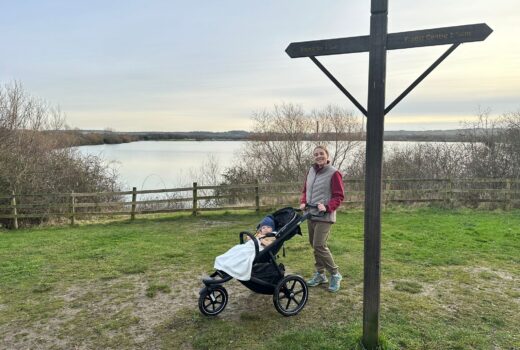 Woman with a running buggy stands on the grass by a wooden sign, with a lake in the background