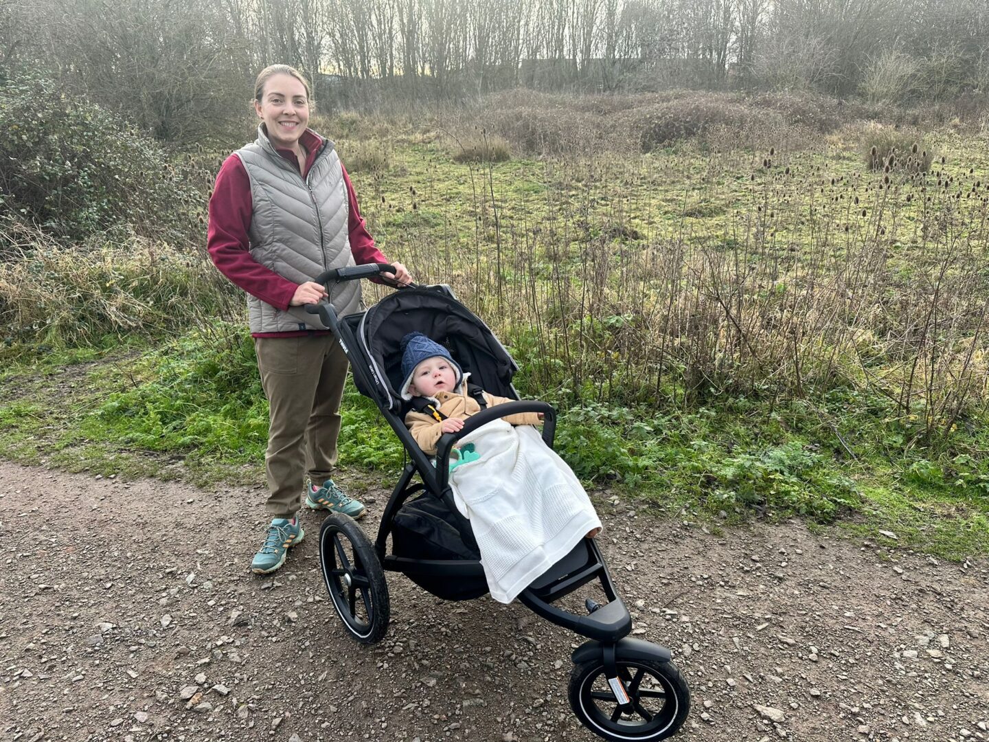 Woman pushing an offroad buggy with a baby in it. They stand on a gravel path with grass and trees behind them. 