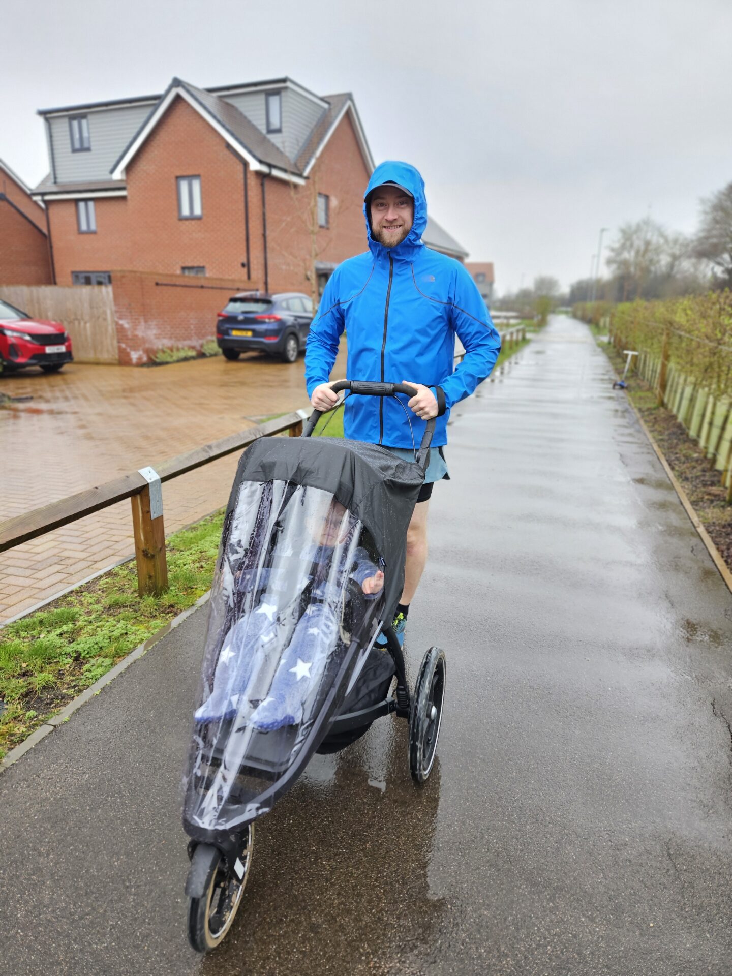 Man wearing blue waterproof coat pushes a running buggy with raincover on 